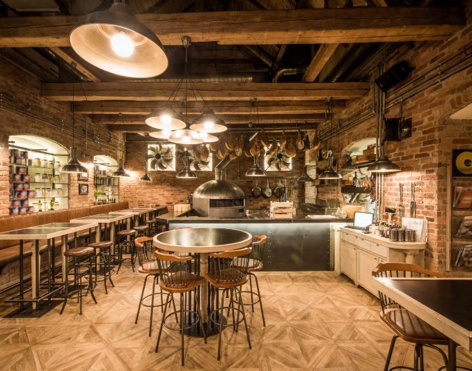 Restaurant interior with exposed brick, wooden beams, round tables, and a pizza oven.