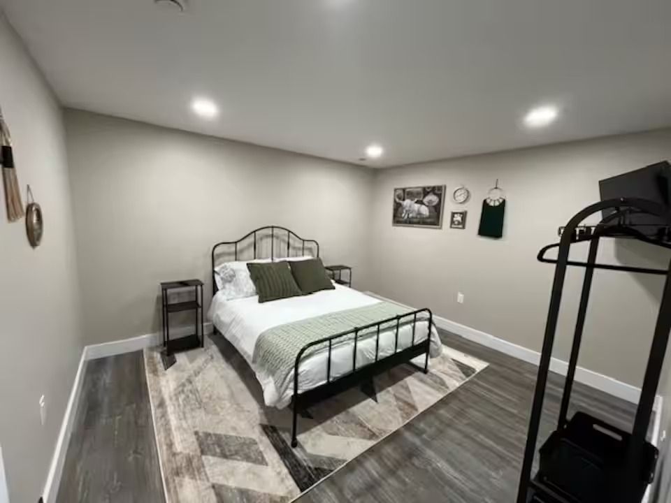 A modern, neutrally toned bedroom featuring a metal-framed bed with green pillows, a patterned rug, and a clothing rack.