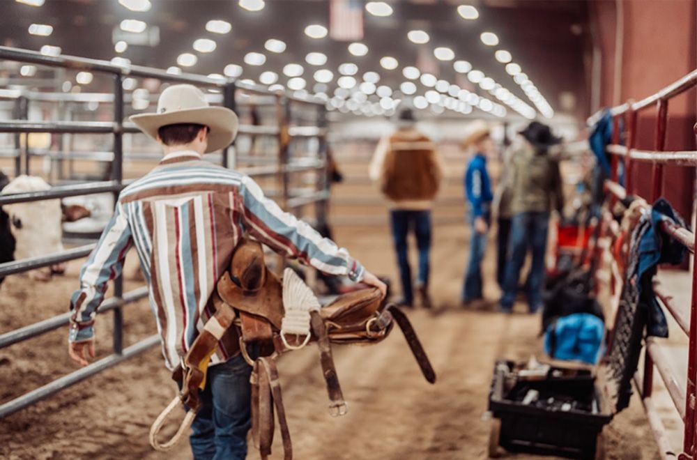 Cowboy carrying saddle in an indoor arena, looking towards other people.
