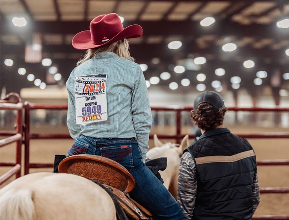 Cowgirl in red hat on horse, watching rodeo event. Man in vest beside her. Indoor arena setting.
