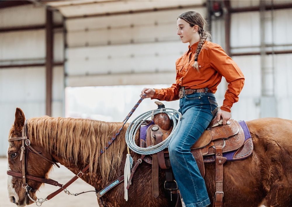 Young person riding a horse in a riding arena, wearing a rust-colored shirt and jeans.