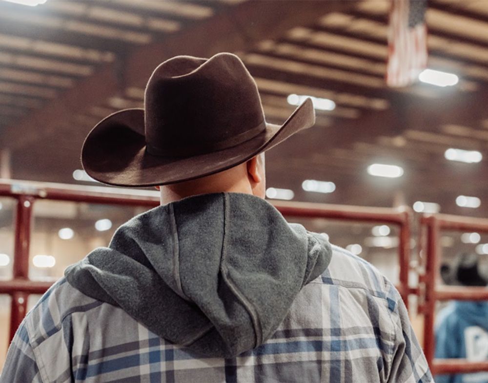 Man in cowboy hat and hooded sweatshirt at rodeo, looking towards arena.
