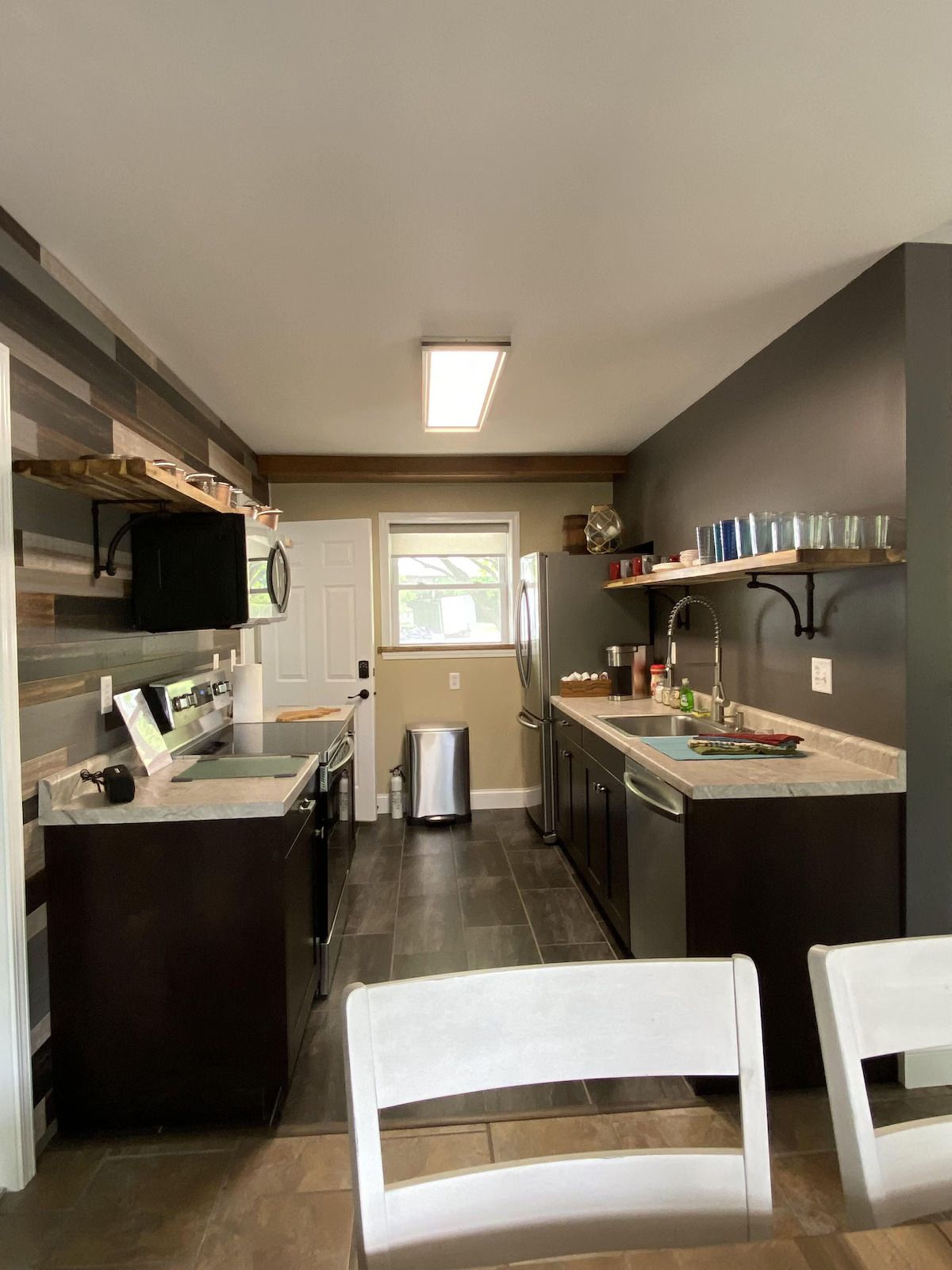 A galley-style kitchen featuring dark wood cabinets, light countertops, open shelving, and a stainless steel trash can.