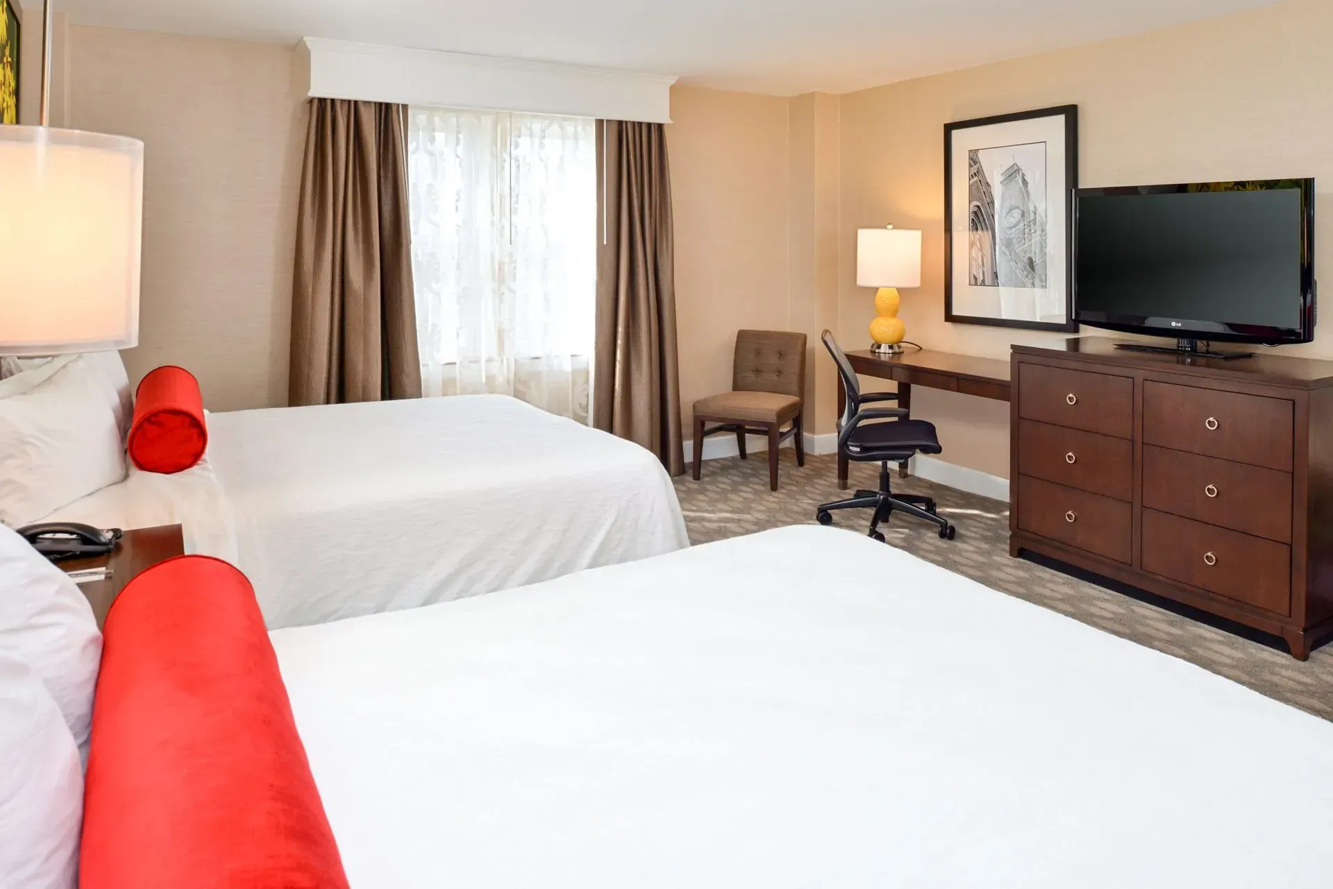 A hotel room with two beds, a desk, a rolling chair, a dresser with a television, and a window with brown curtains.