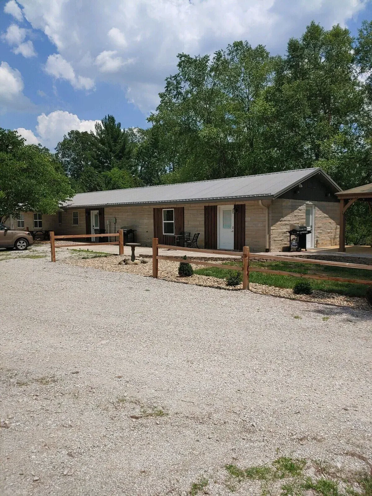 A single-story, light-colored brick building with a gray metal roof, surrounded by trees and a gravel parking lot.