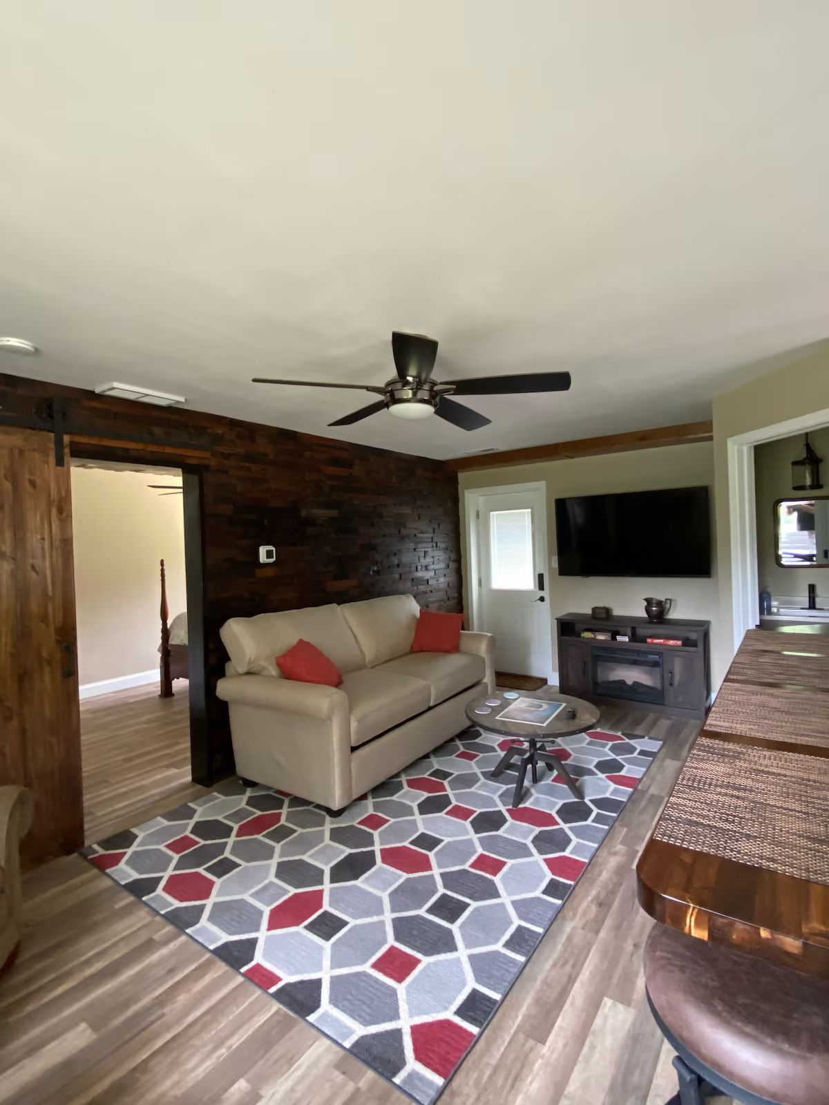 Living room with a beige sofa, geometric rug, rustic wood wall, ceiling fan, and television above a media console.