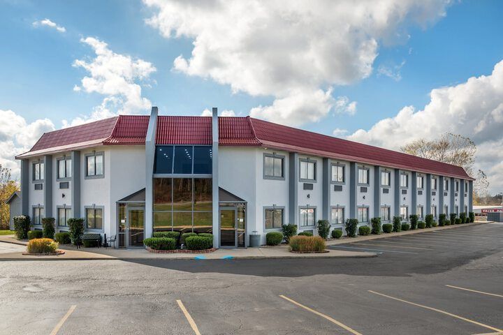 Two-story white hotel with red roof and blue glass entrance, viewed from a parking lot under a partly cloudy sky