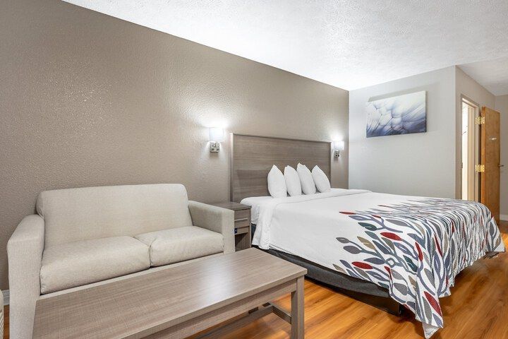 Hotel room with bed, sofa, nightstand, wall lamp, and wooden floor under soft lighting