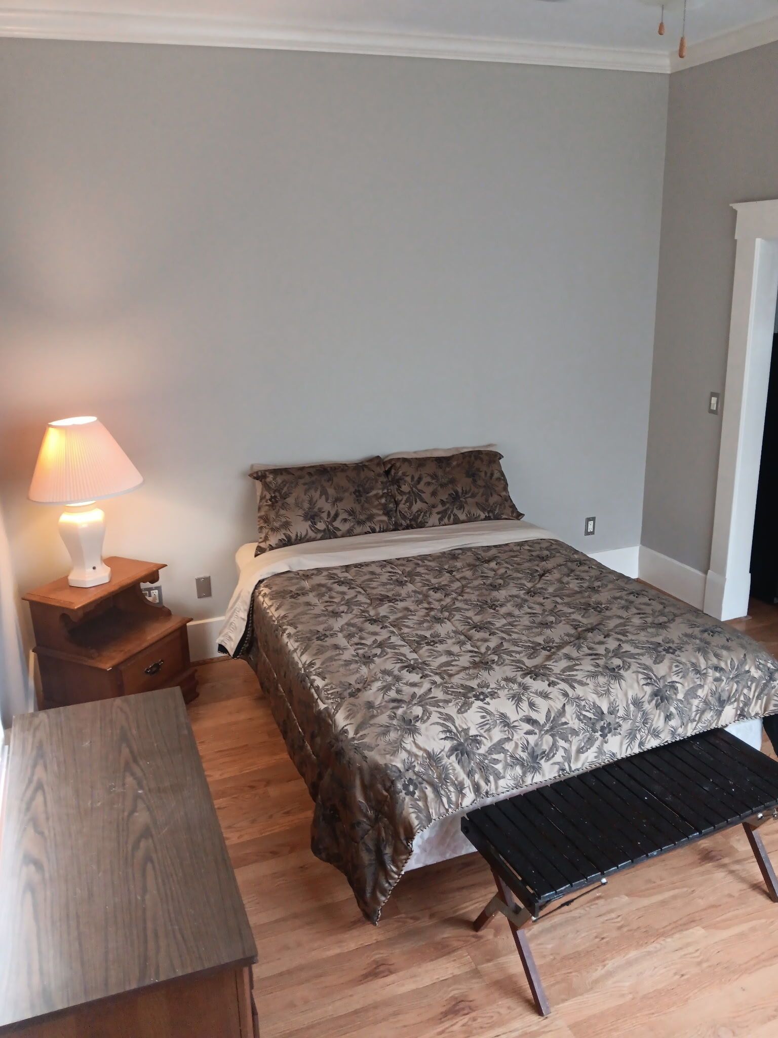 Bedroom with a bed, nightstand, and bench. Gray walls, patterned comforter, wood floors, and natural light.