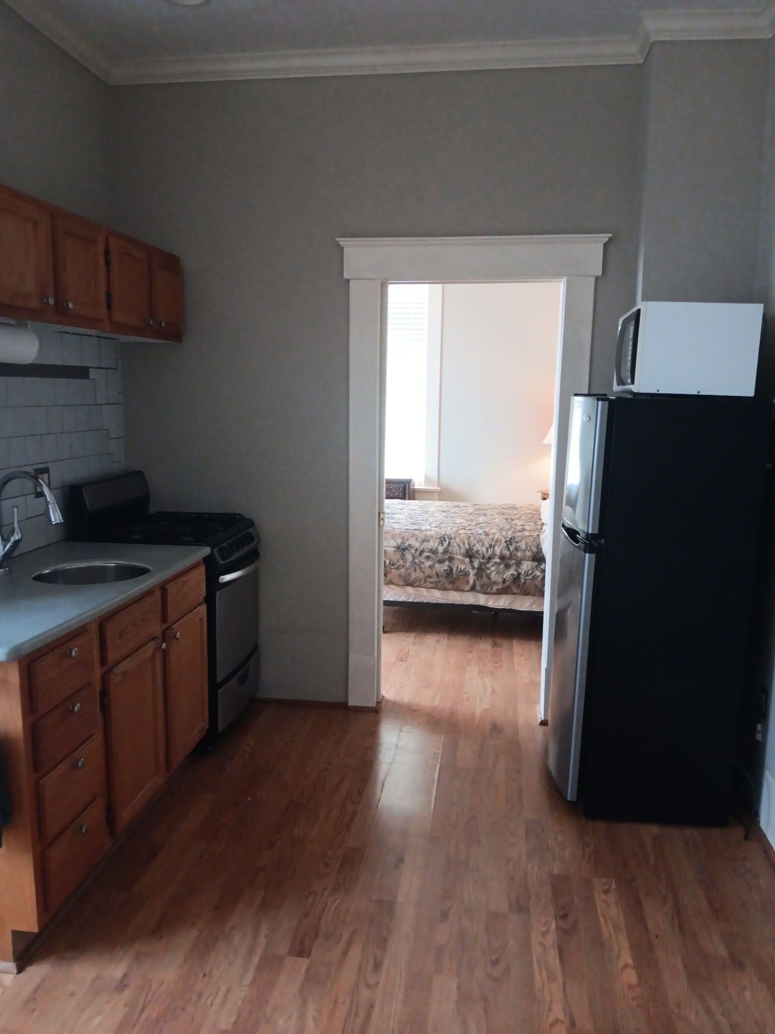 Kitchen with wood cabinets, gray walls, and a doorway leading to a bedroom. Black refrigerator on the right.