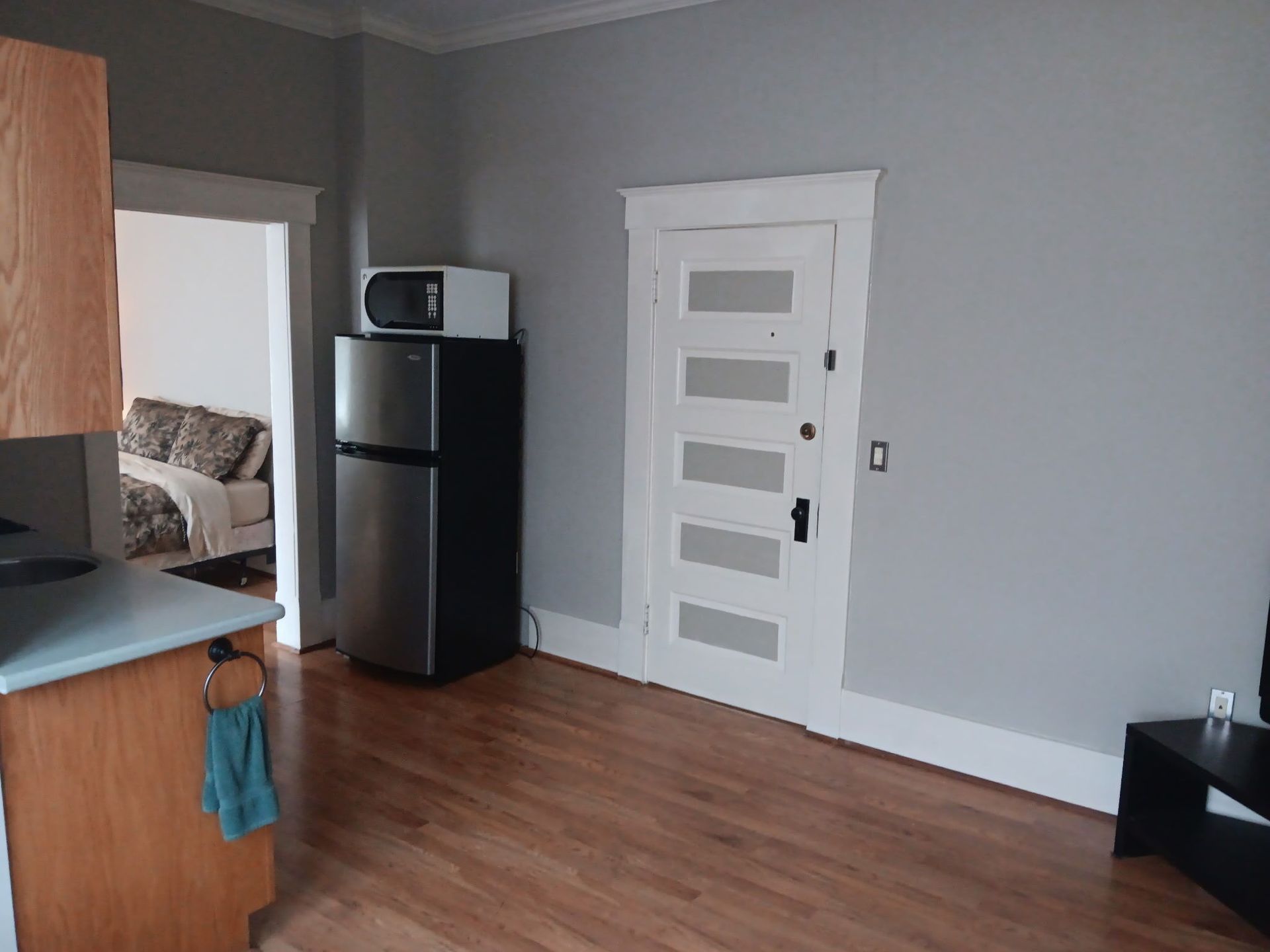 A small kitchen and doorway with hardwood floors. Refrigerator, microwave, and a view into a bedroom.