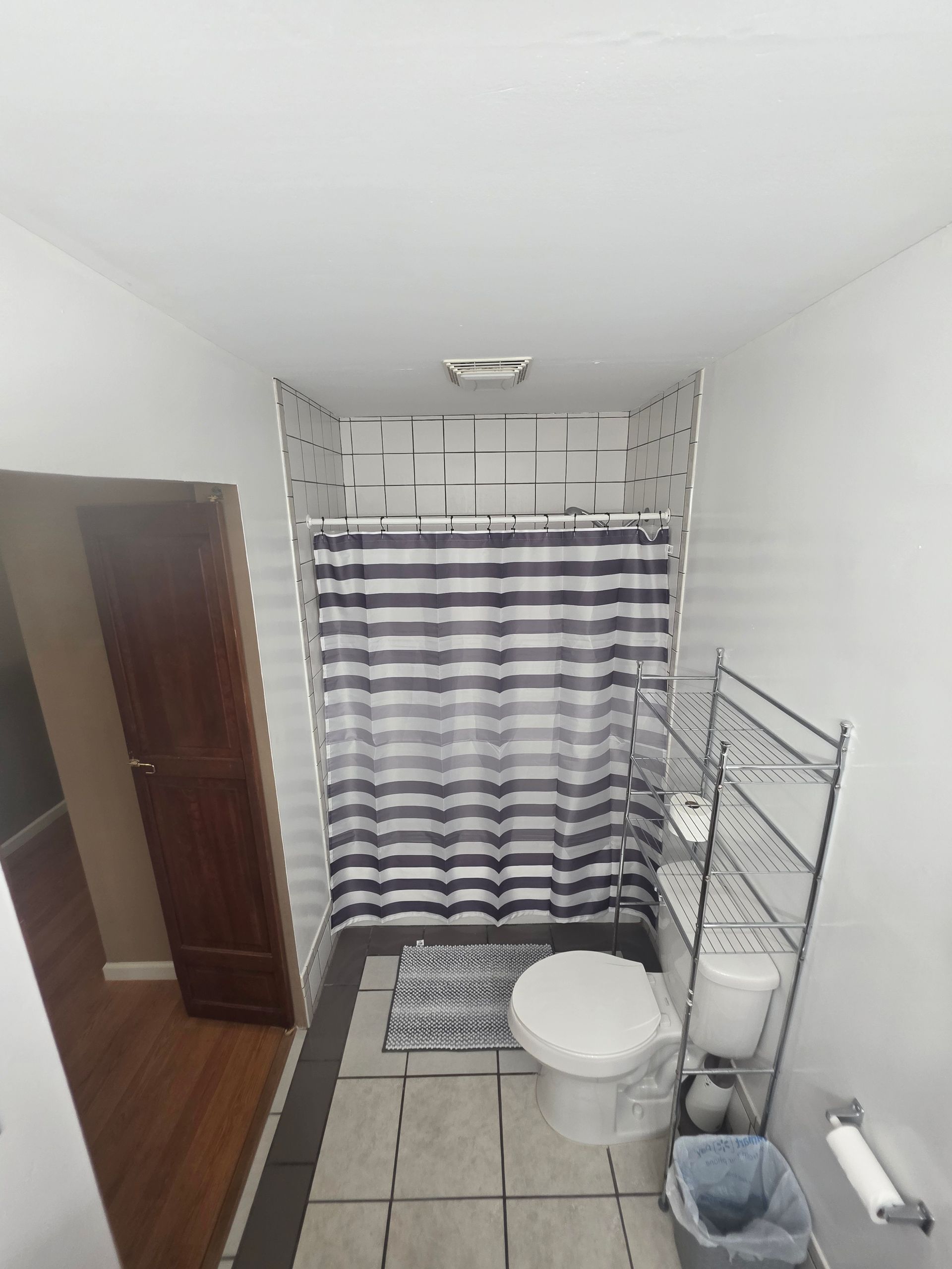 Bathroom with toilet, shower, and shelving. Black and white shower curtain. Gray and white tiled floor.