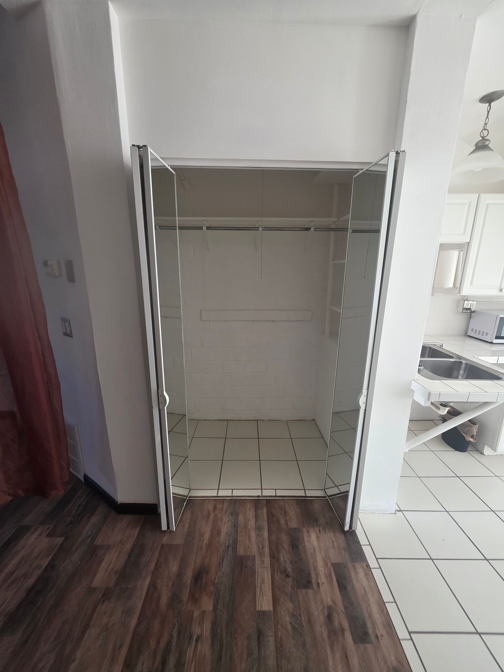 An empty, white-walled closet with open doors on a wood-look floor, near a kitchen.