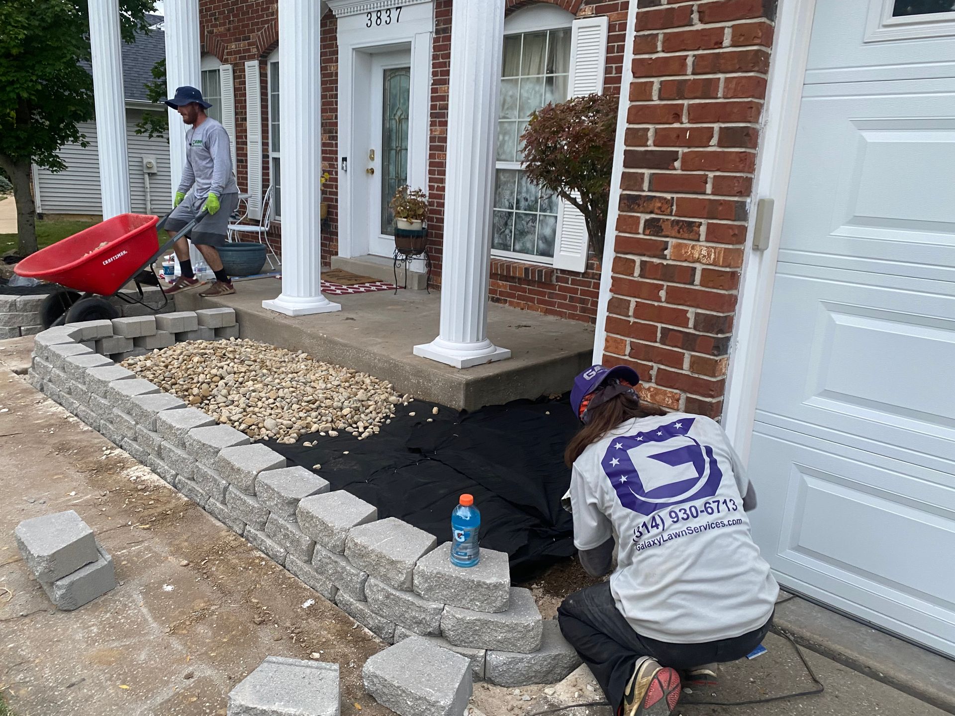 Workers landscaping a front porch with stone edging and gravel at a brick house.