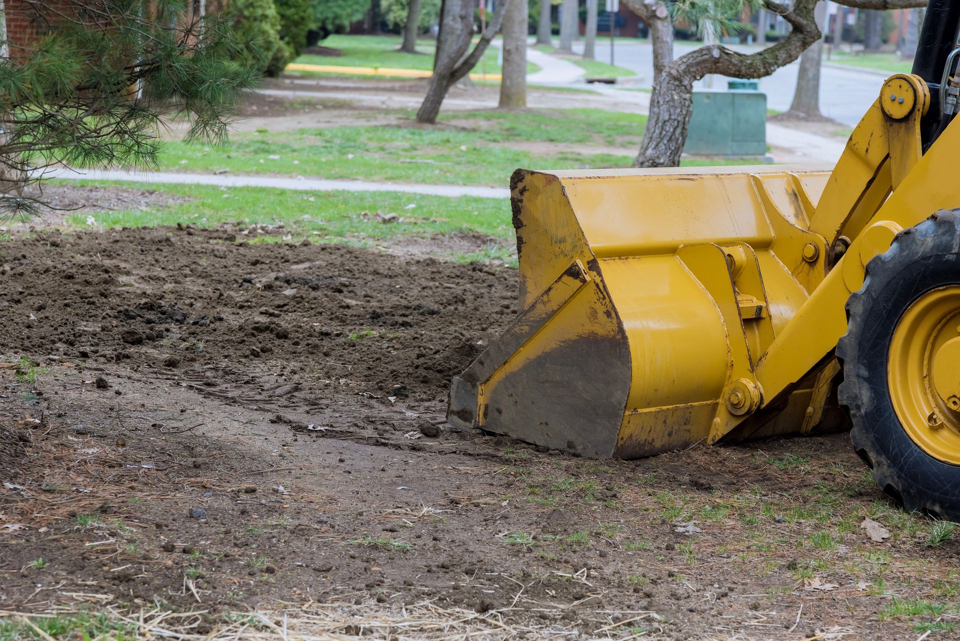 A yellow front-loader machine levels dark soil on a grassy lawn in a residential neighborhood.