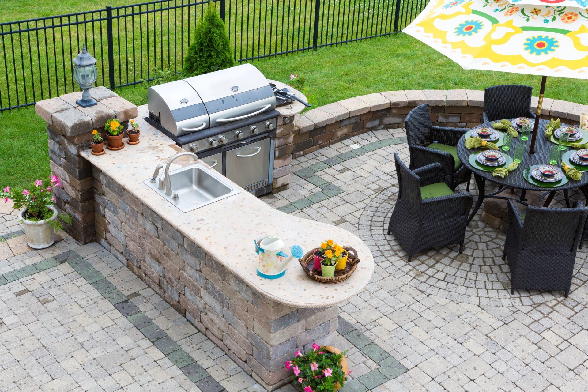 A backyard patio with a stone outdoor kitchen, sink, grill, and a round dining table with chairs under an umbrella.