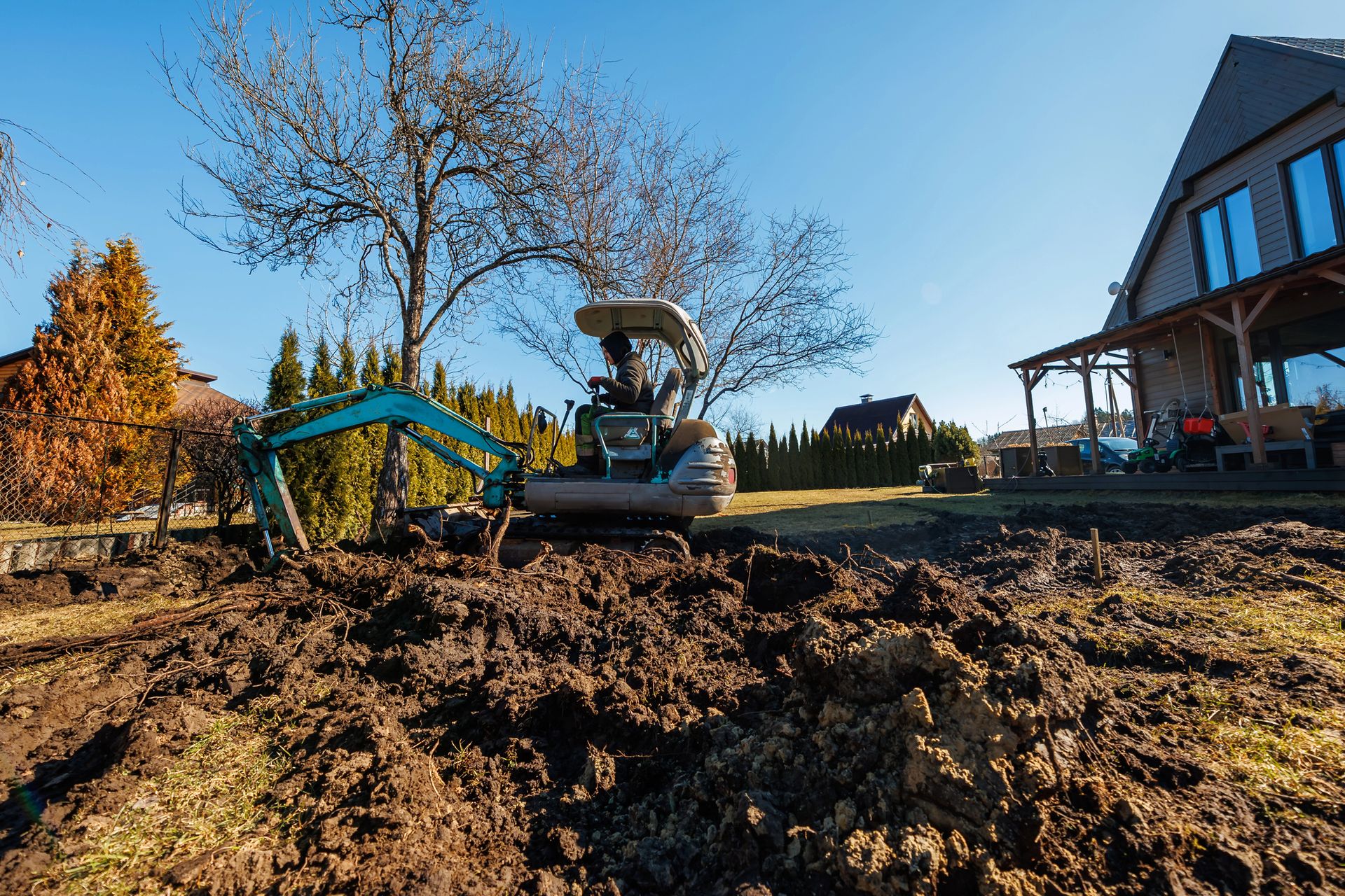 Excavator and tractor working in a muddy yard beside a house under a blue sky