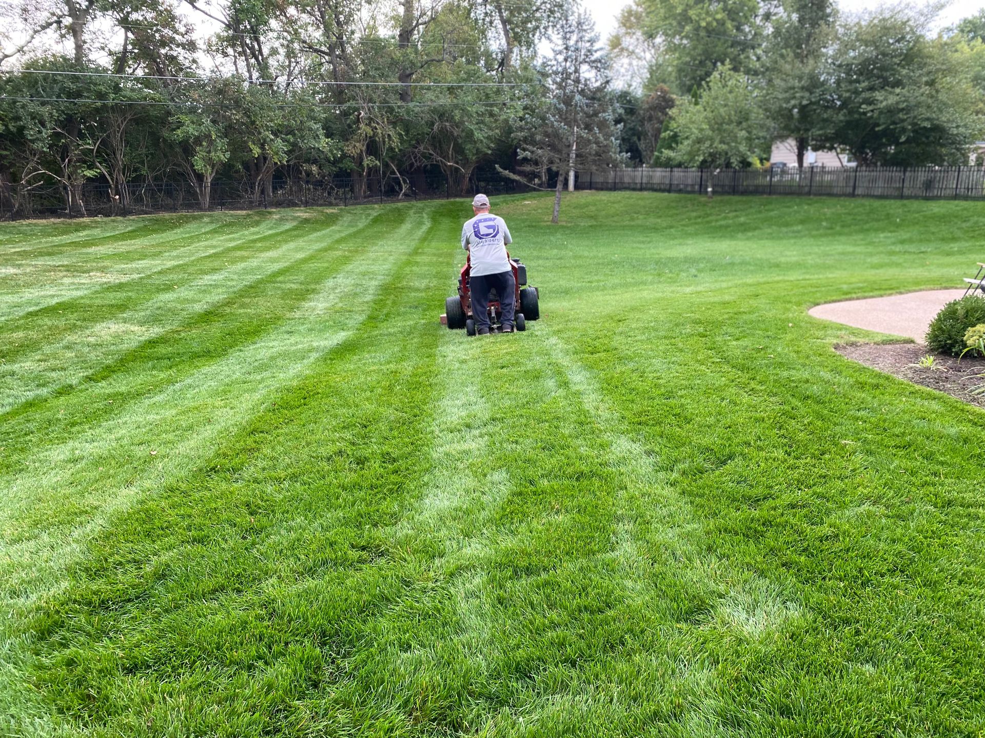 Person mowing a large lawn, creating striped patterns on the green grass. Trees and a house in the background.