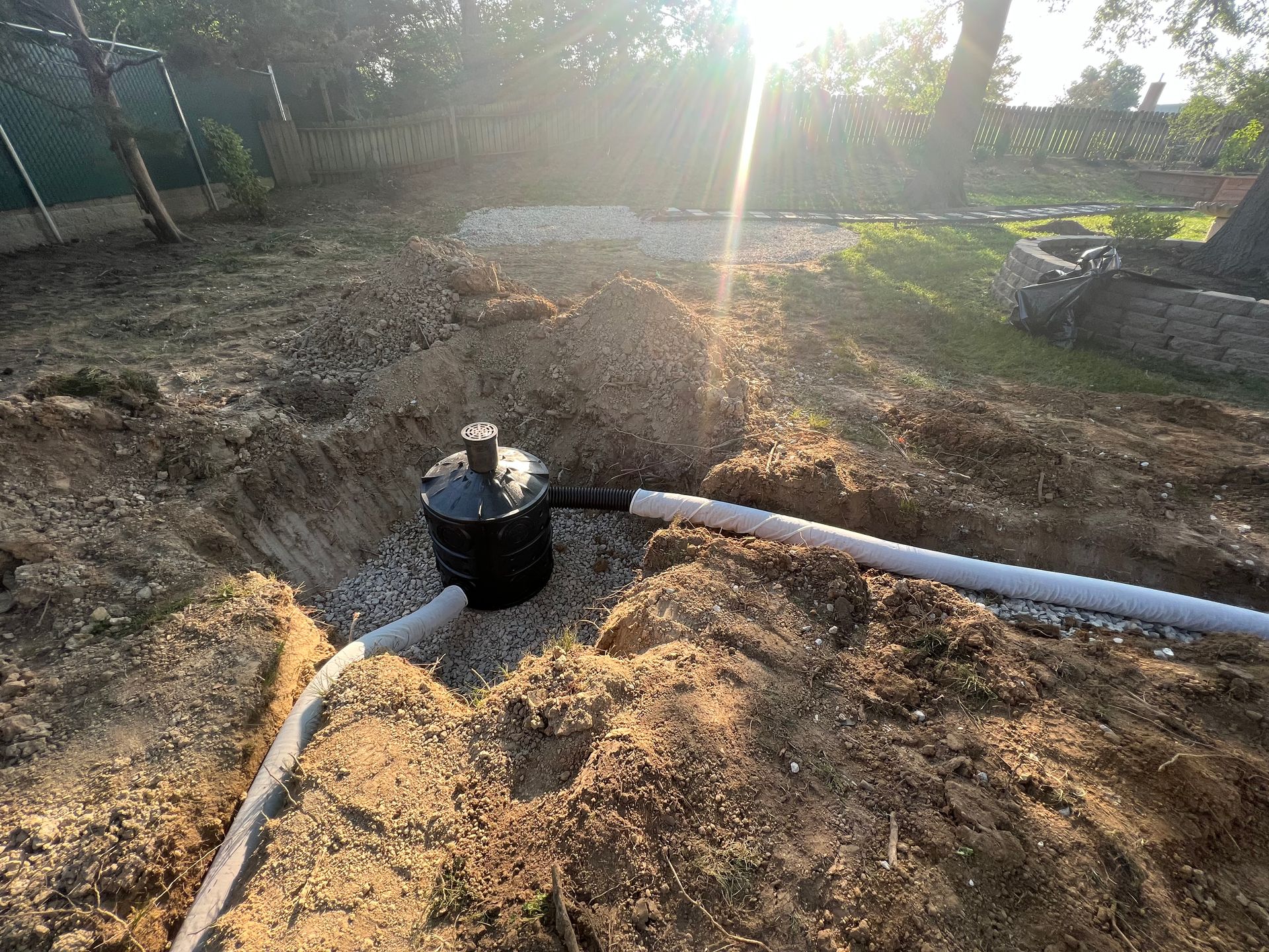 A black drainage basin installed in an excavated pit in a backyard, connected to white drainage pipes surrounded by gravel.