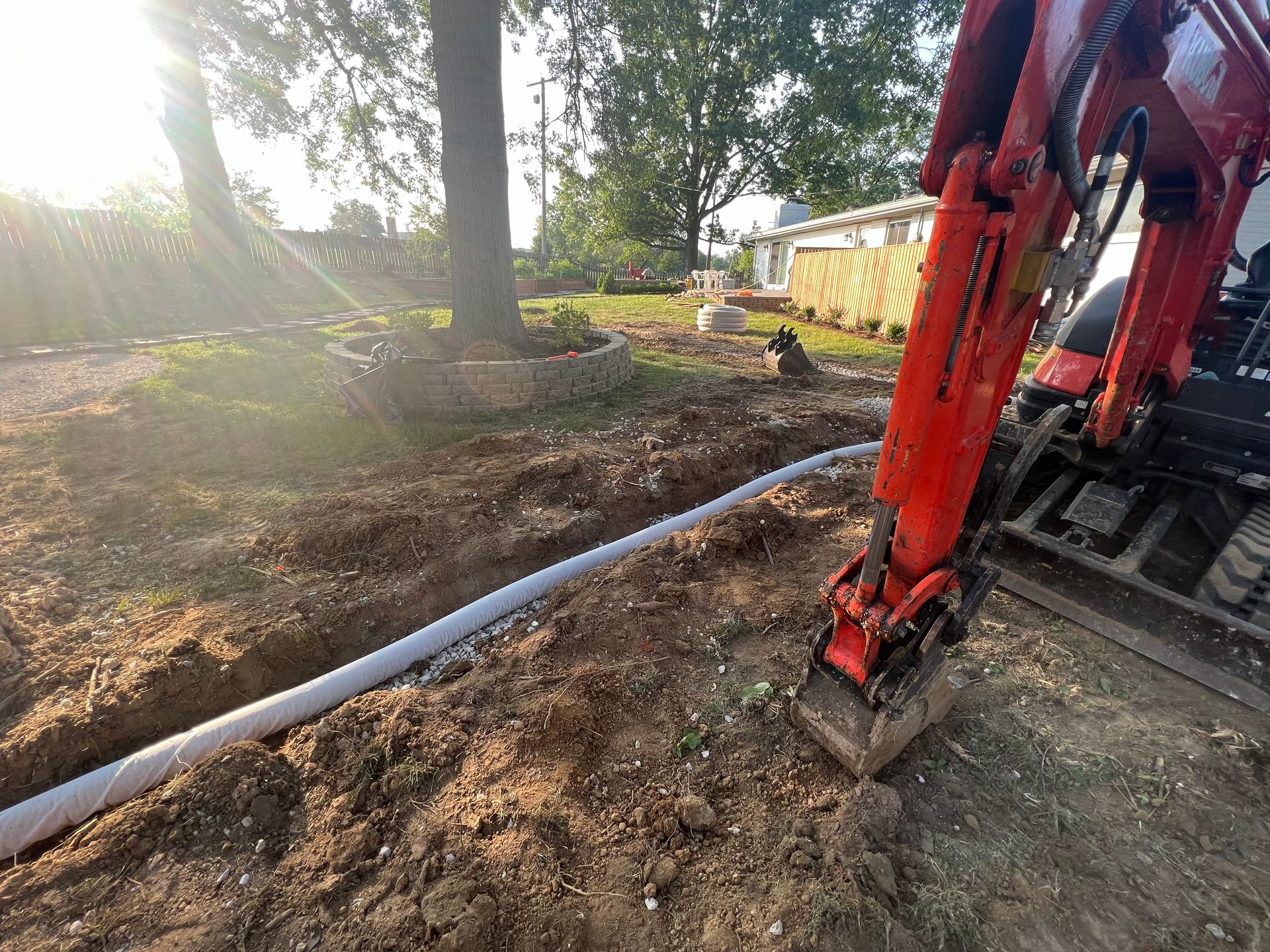 Mini excavator laying drainage pipe in a backyard. Earth tones with green grass and trees.