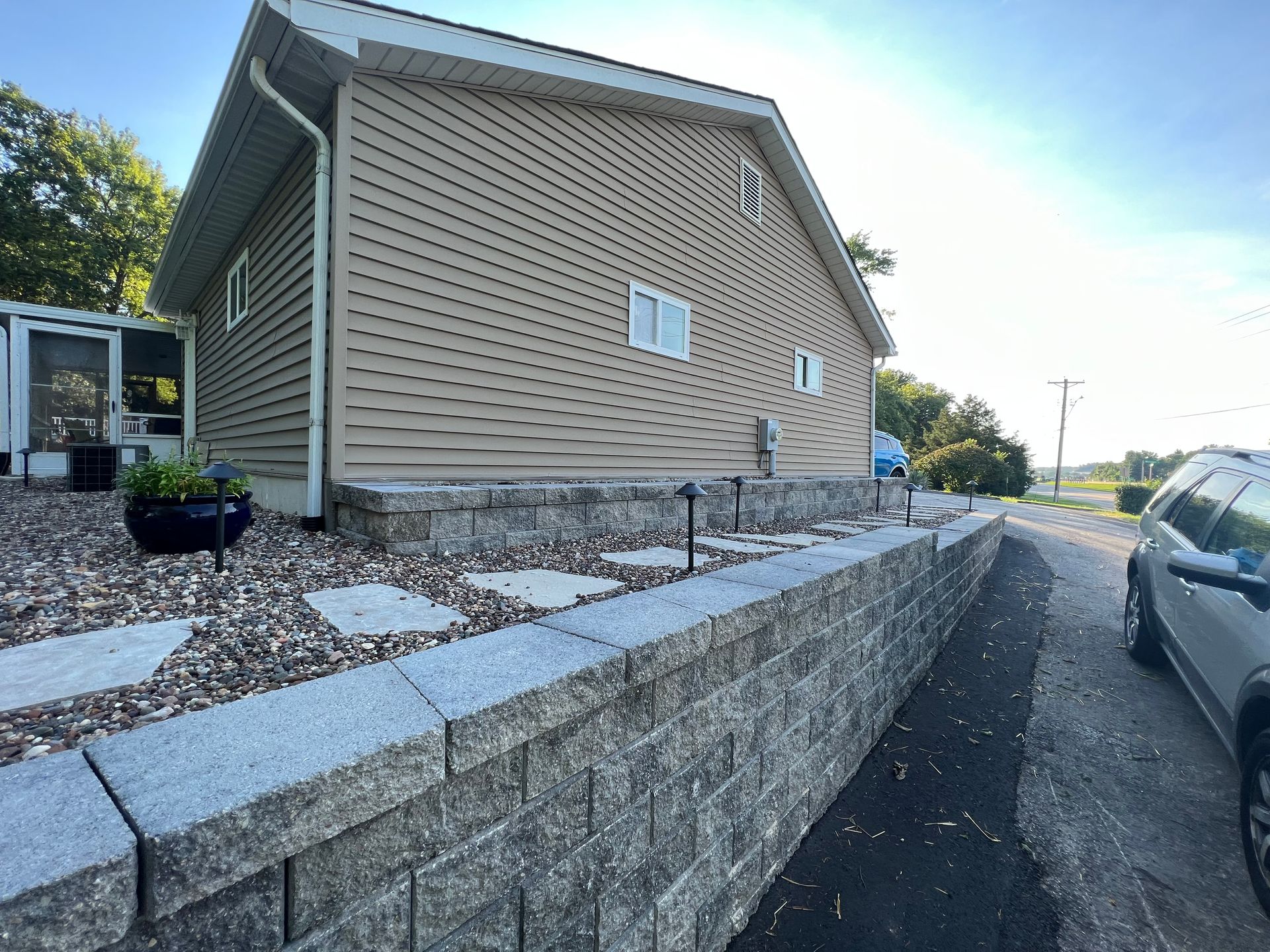 A tan-sided house with a large stone retaining wall and stone path running alongside a driveway on a sunny day.