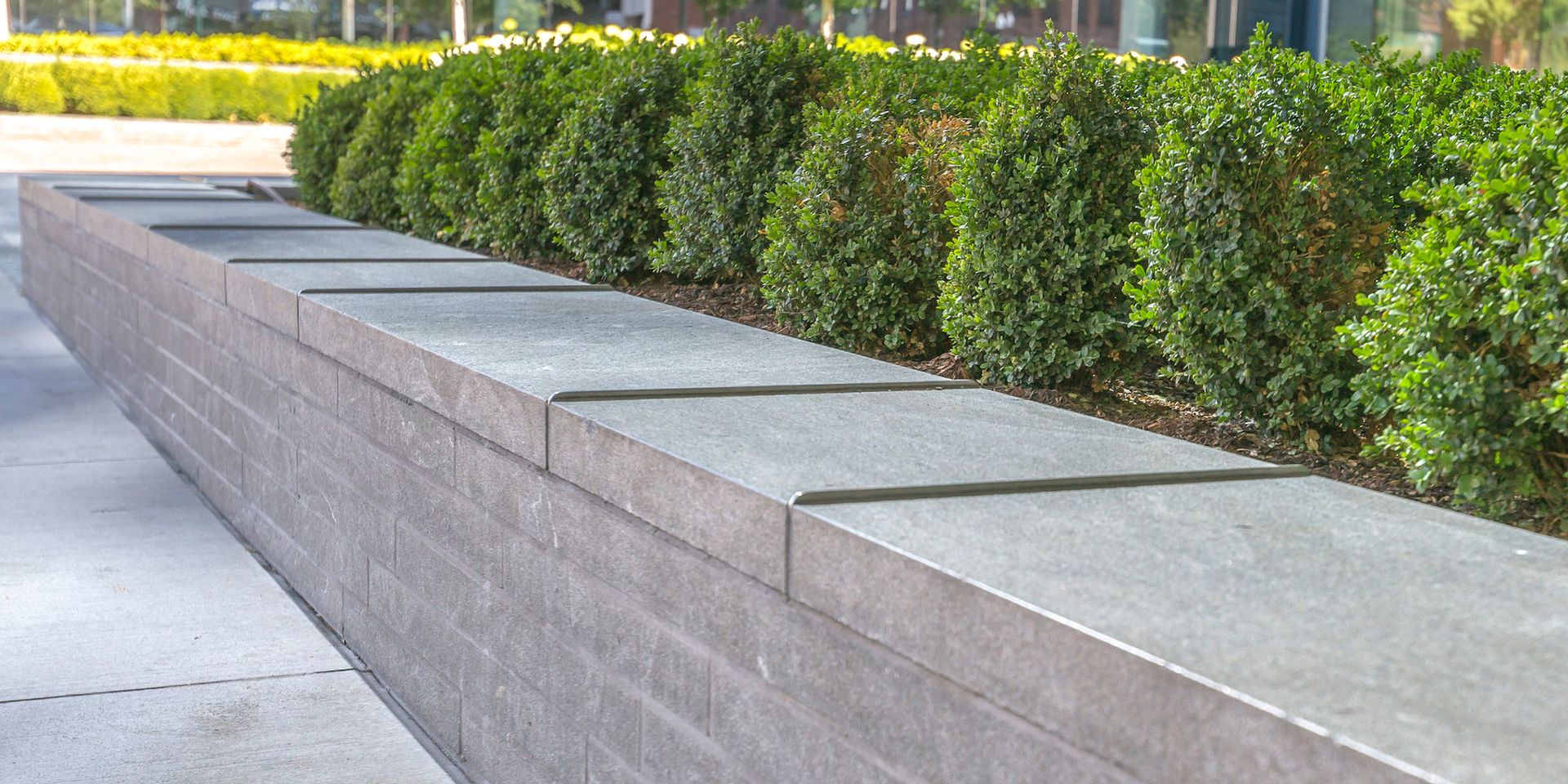 A gray brick retaining wall with flat stone capping, fronting a row of green bushes on a sunny day.