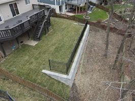 Backyard with a deck, retaining wall, and lawn bordered by a fence and trees.