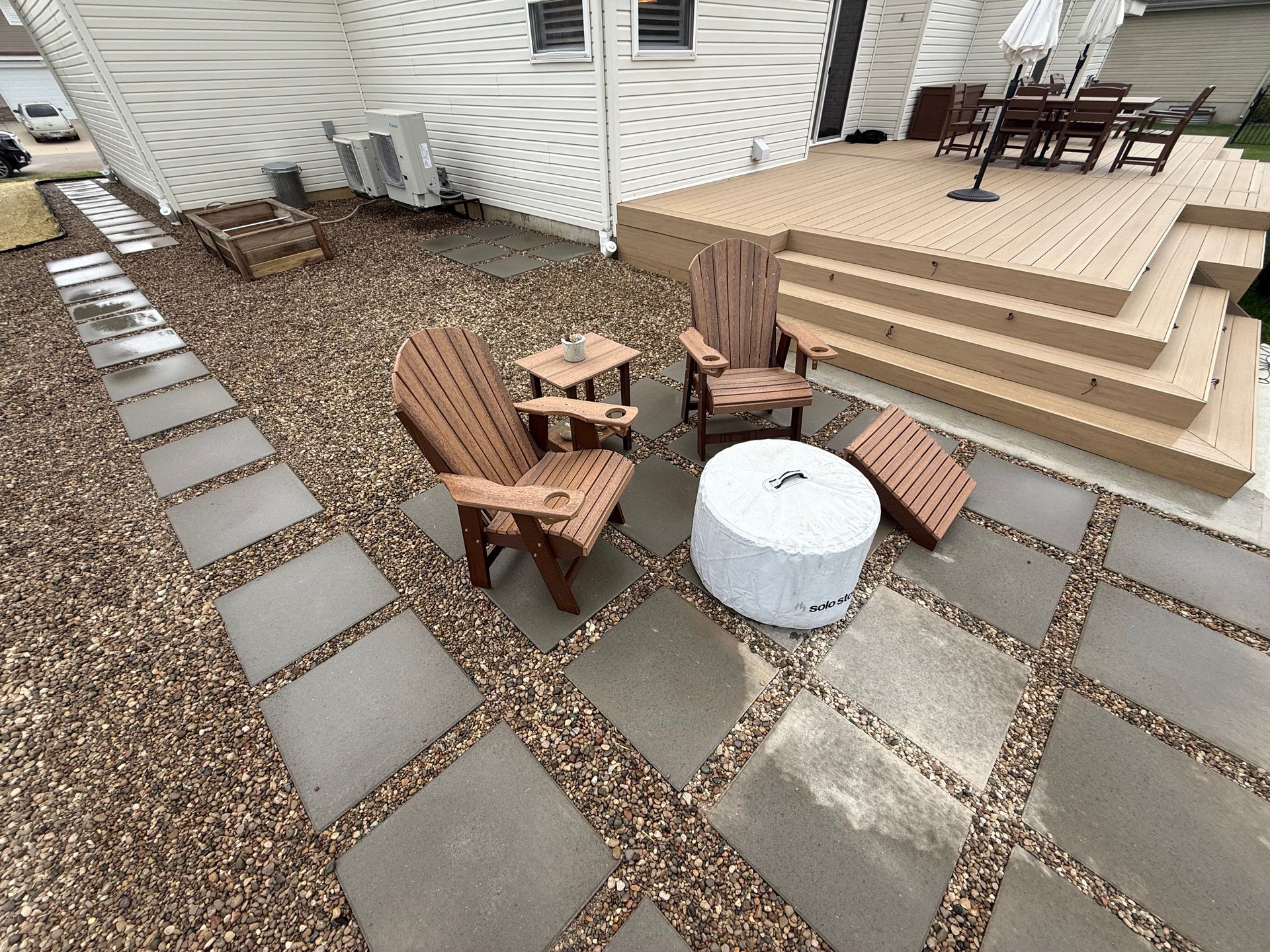 Backyard patio with chairs around a fire pit, stone pavers, and a raised wooden deck with steps.