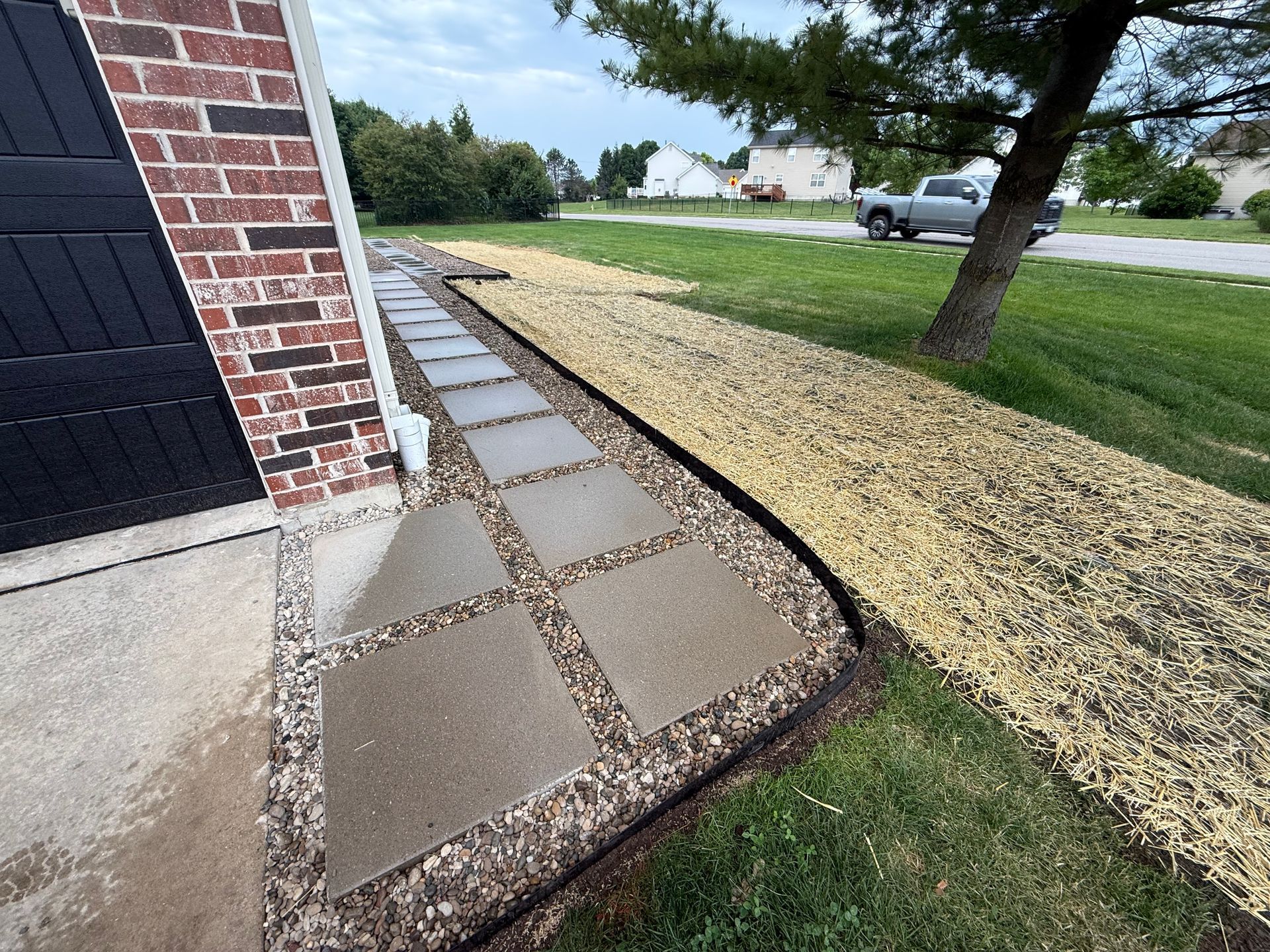 Brick house corner with concrete walkway, mulch border, and grass lawn beside a driveway