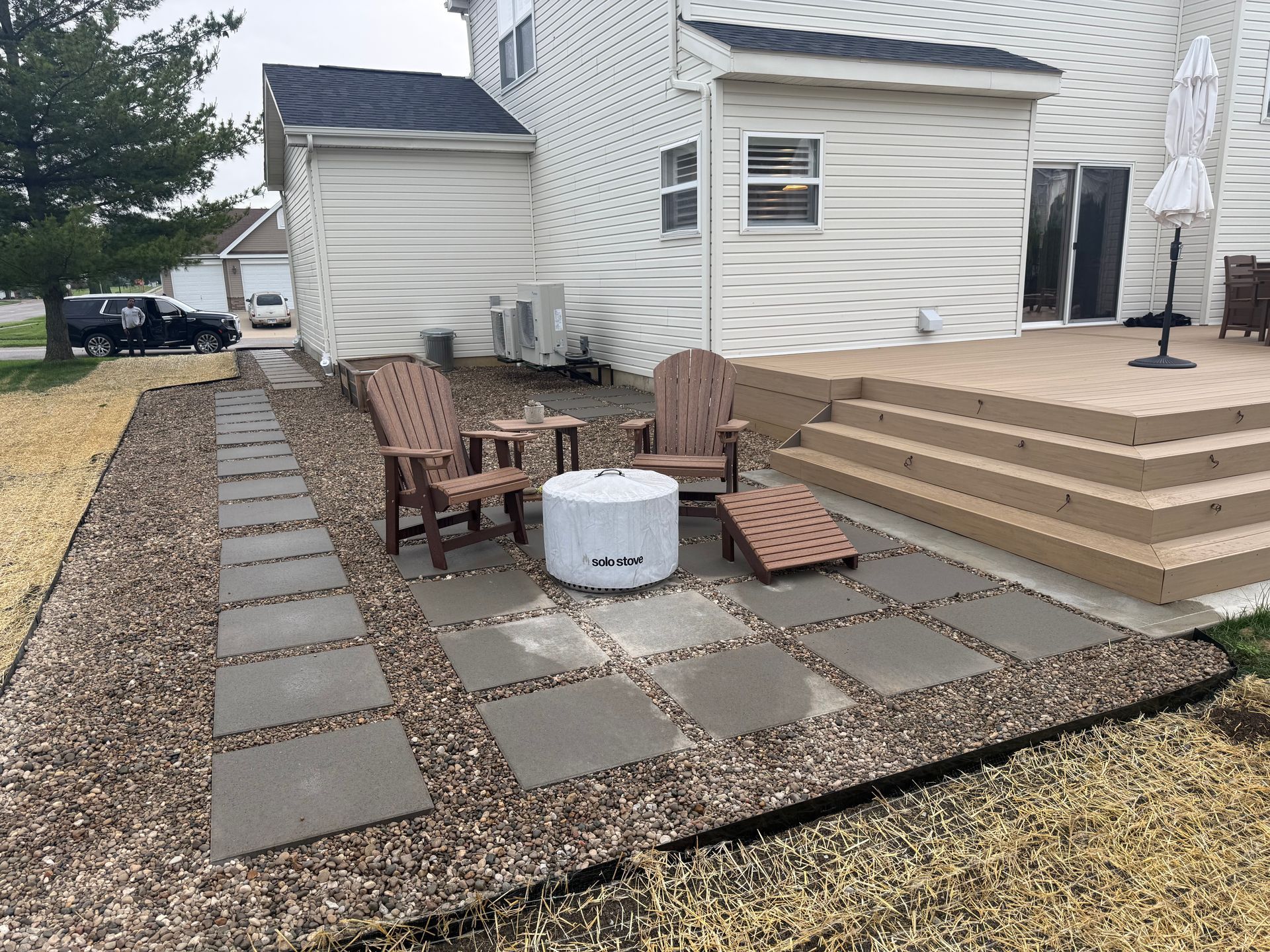 Backyard patio with stone pavers, Adirondack chairs, fire pit, and raised wooden deck beside a house
