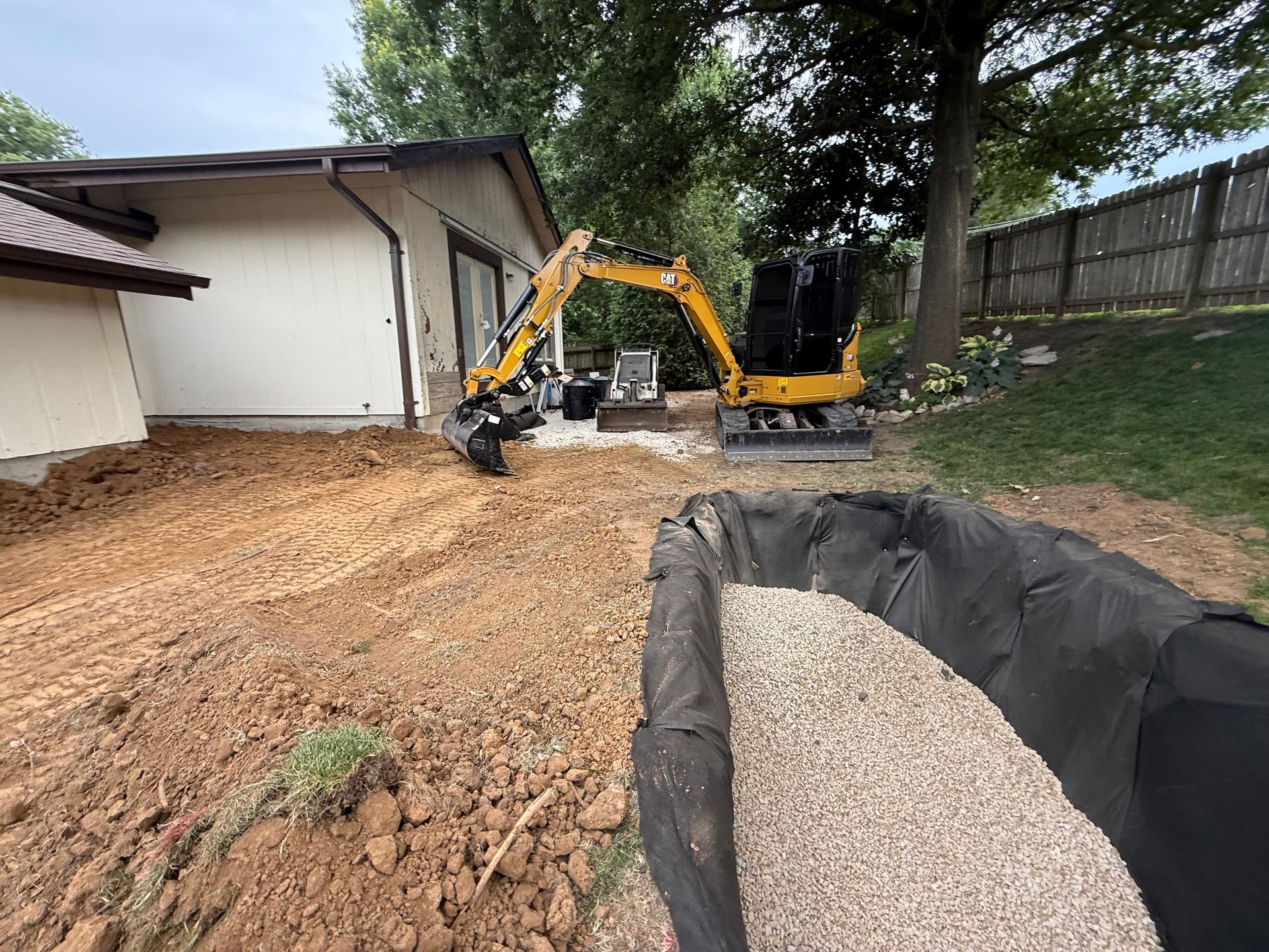 Excavator leveling a dirt yard beside a house, with gravel and a fence in the foreground.