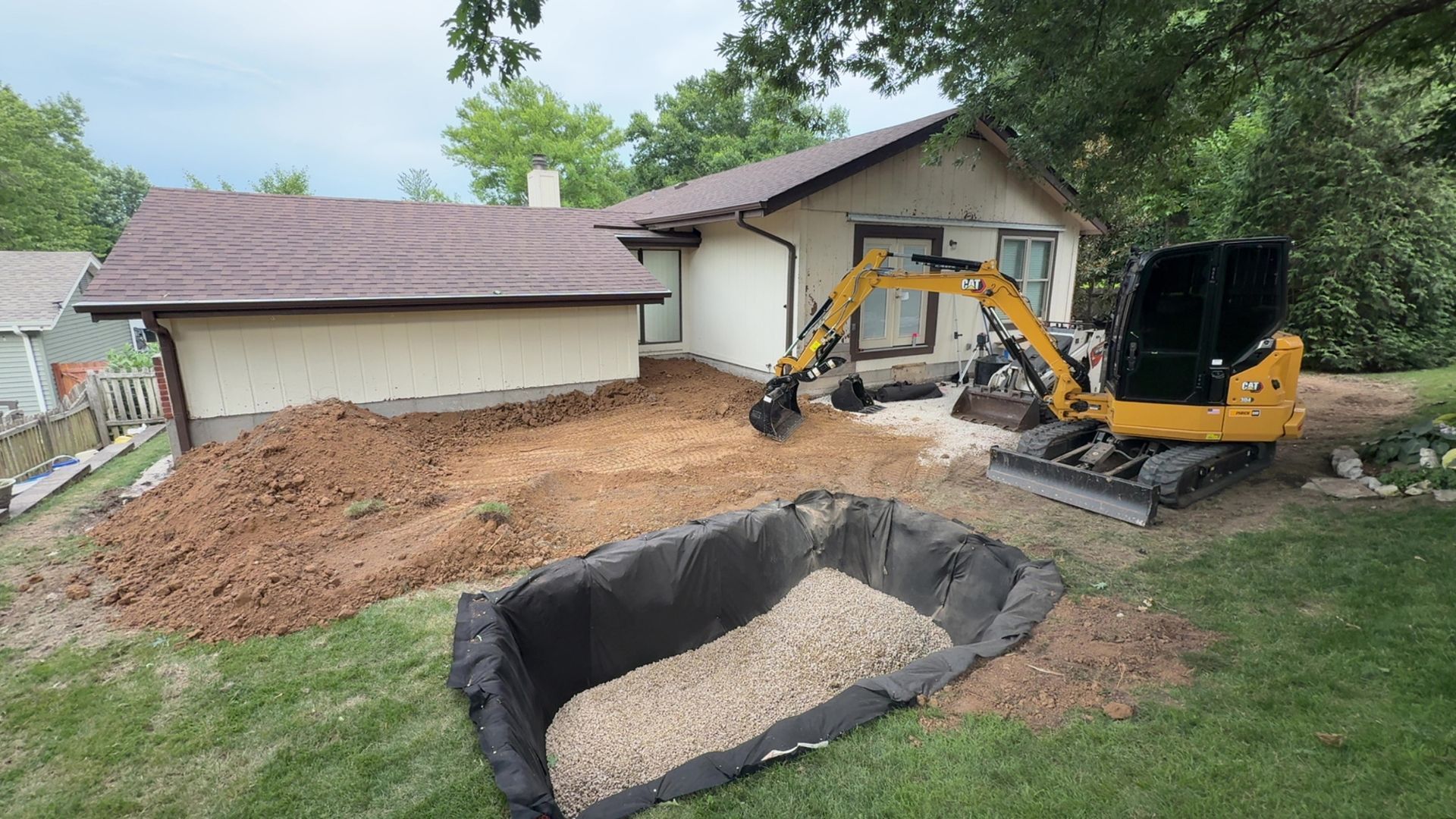 Small excavator at a house digging a large hole beside a tarp-covered pile of dirt