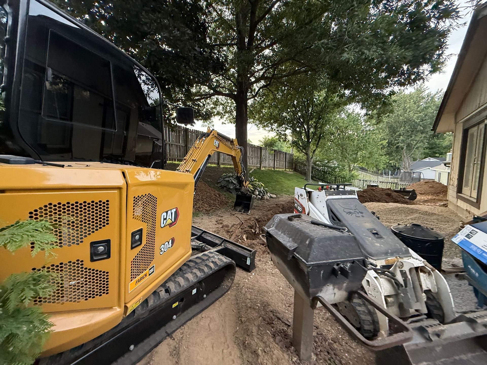Yellow excavator and construction equipment on a muddy path beside a building and trees