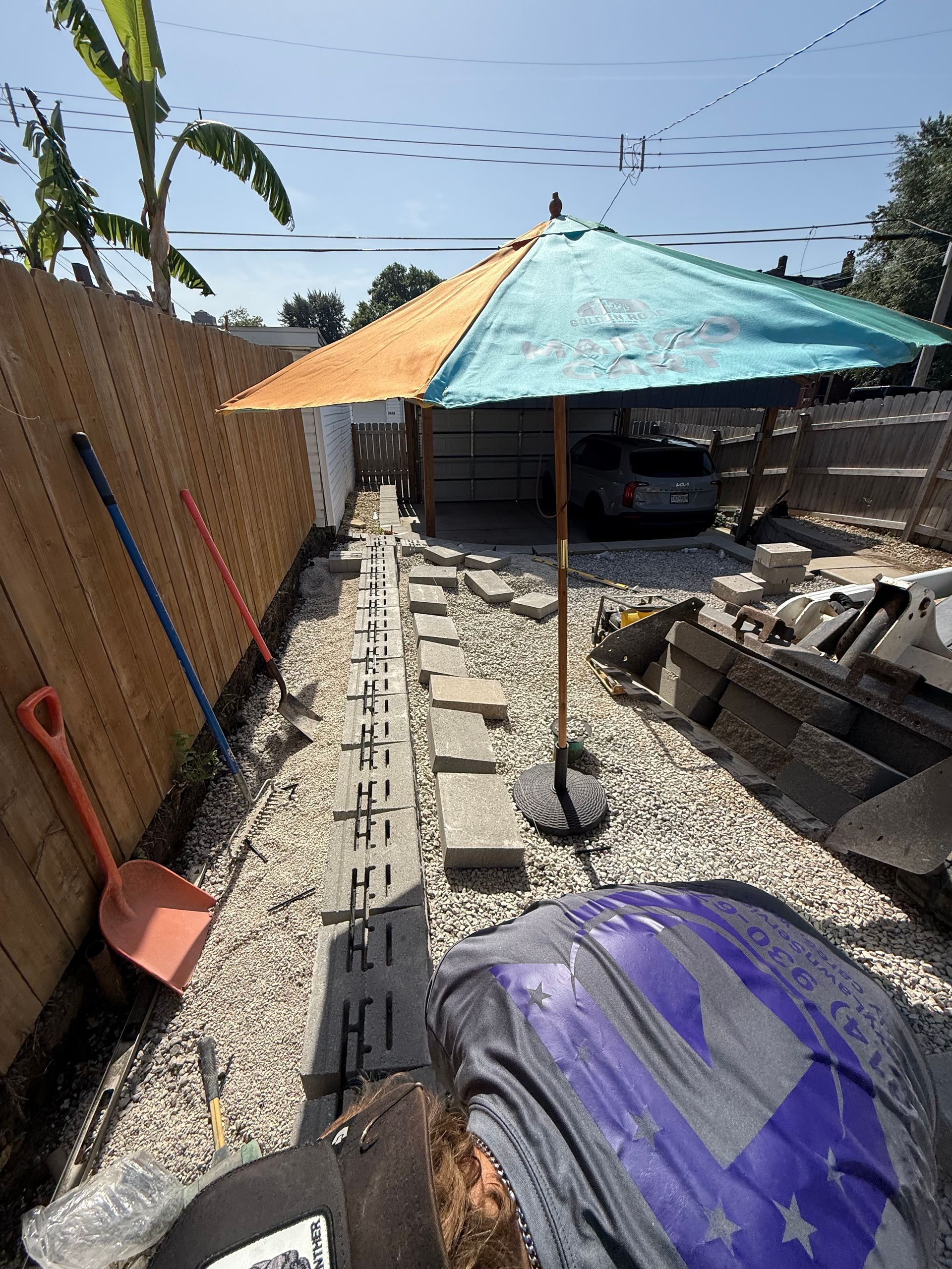 Outdoor gravel patio with a colorful umbrella, cinder blocks, and a fence under a sunny sky