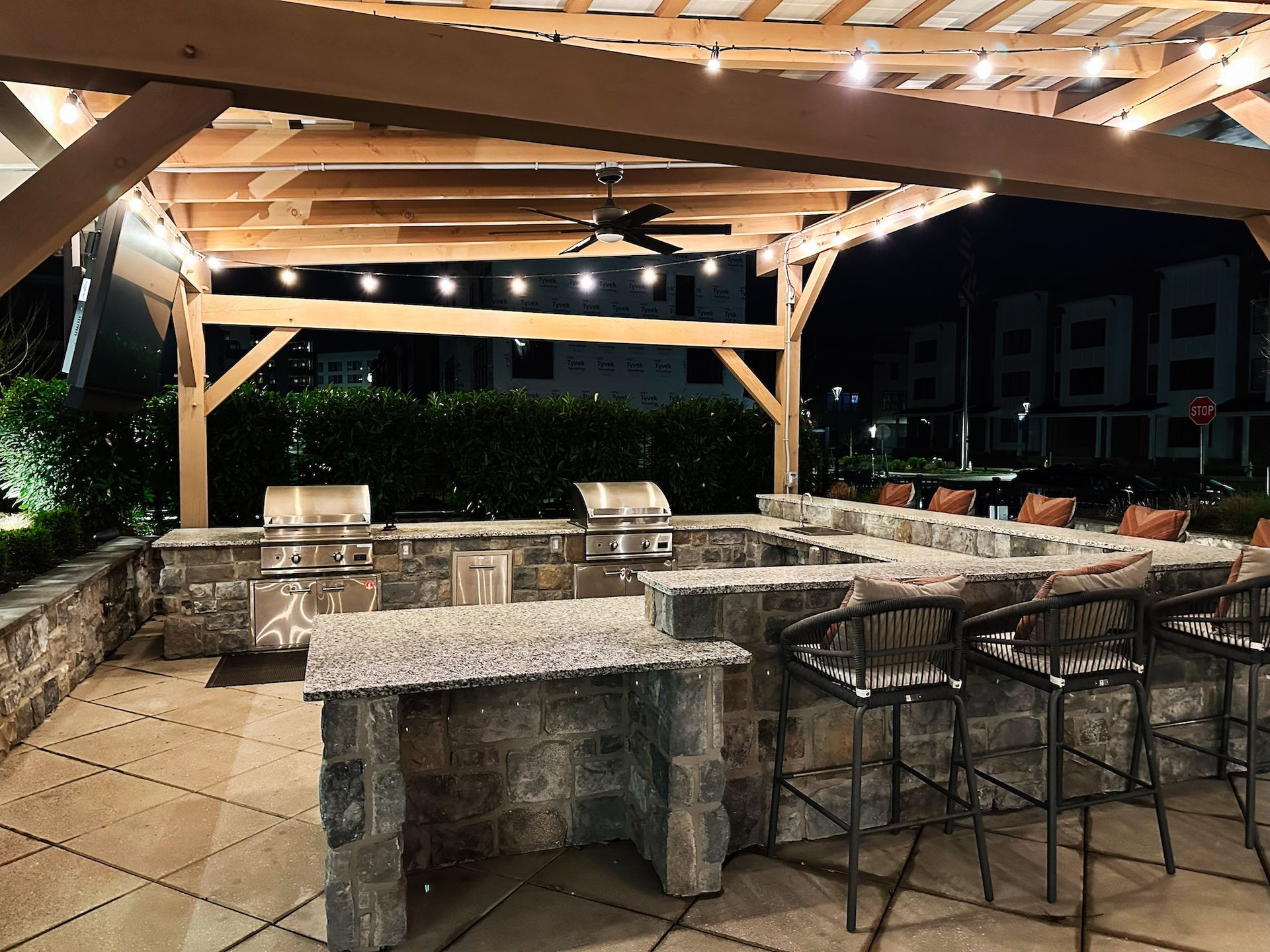 Outdoor kitchen area at night with a stone bar, metal stools, built-in grills, and wood-framed pergola with string lights.