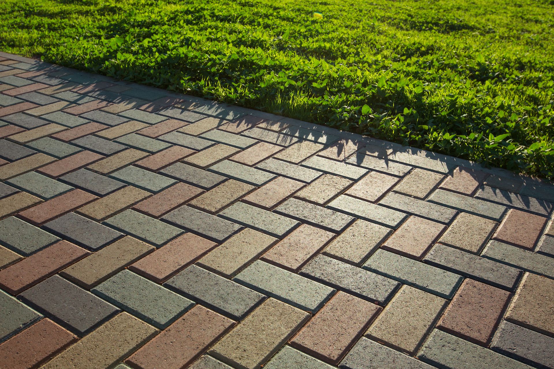 A paved walkway with a herringbone pattern in earthy tones of red, grey, and tan, bordered by vibrant green grass.