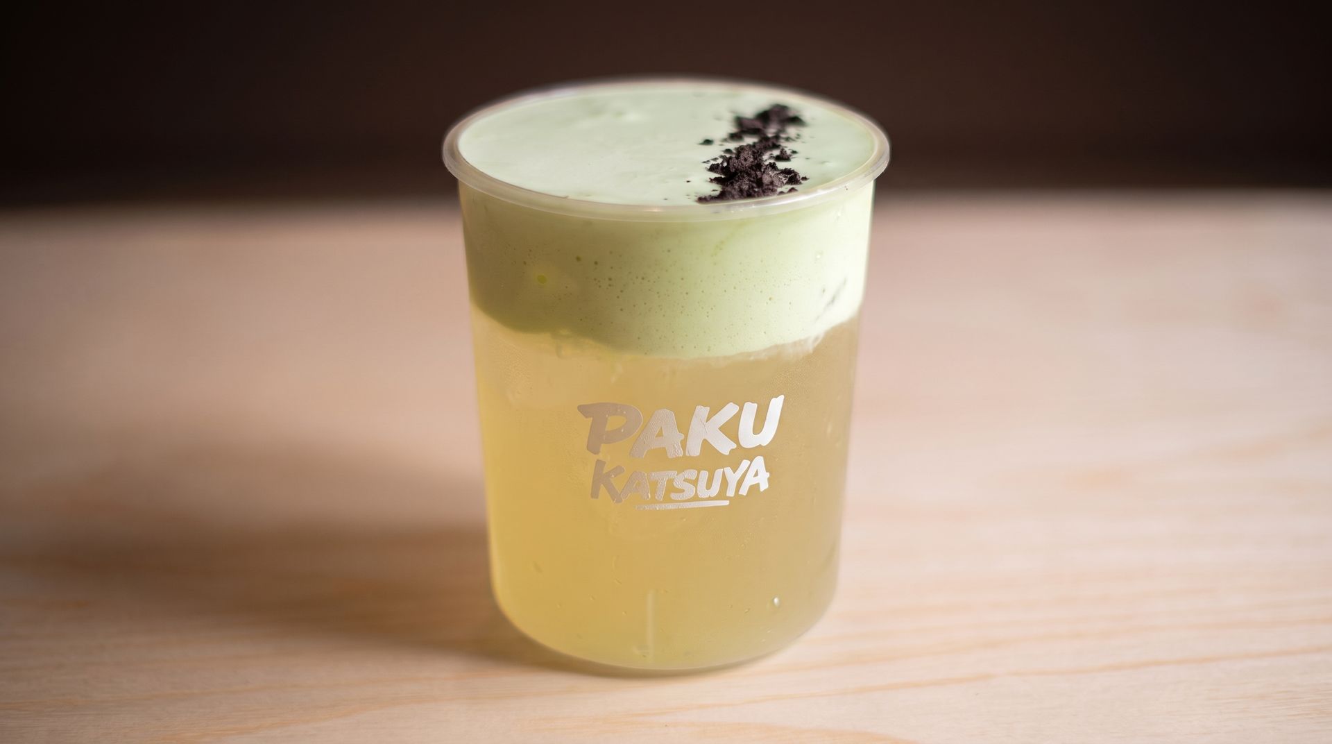 A pale yellow drink in a clear plastic cup with foam and dark topping on a wooden table.