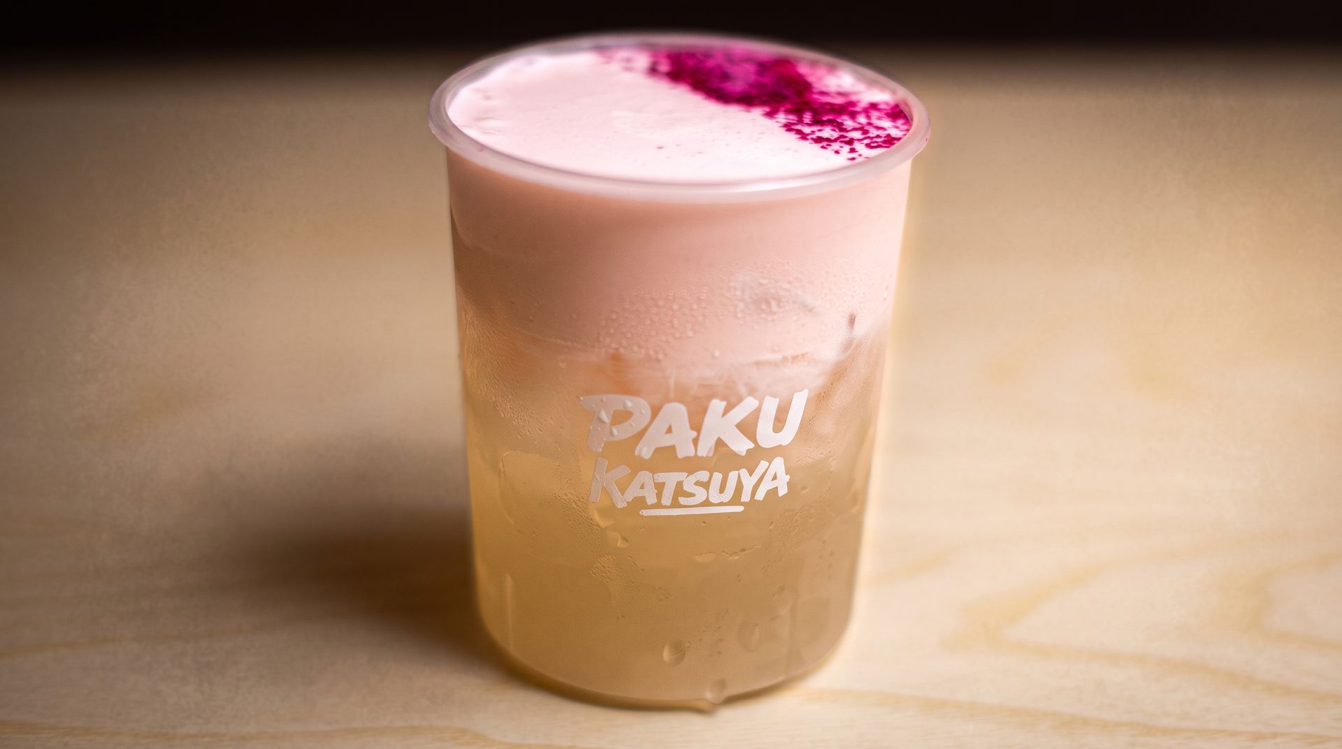 A clear cup of light brown drink topped with pink foam on a wooden table, with PAKU Katsuya logo.