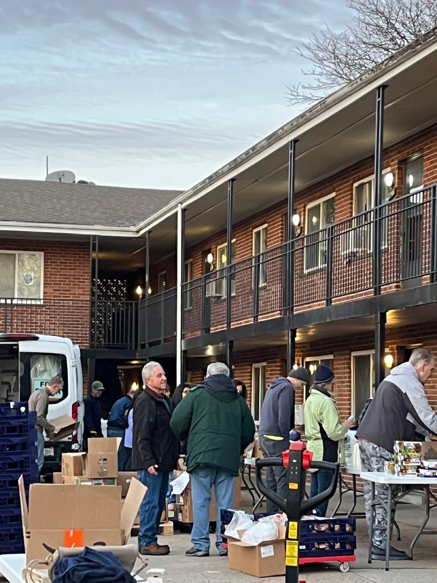 People gathered with boxes and equipment outside a two-story brick building with an upstairs balcony.