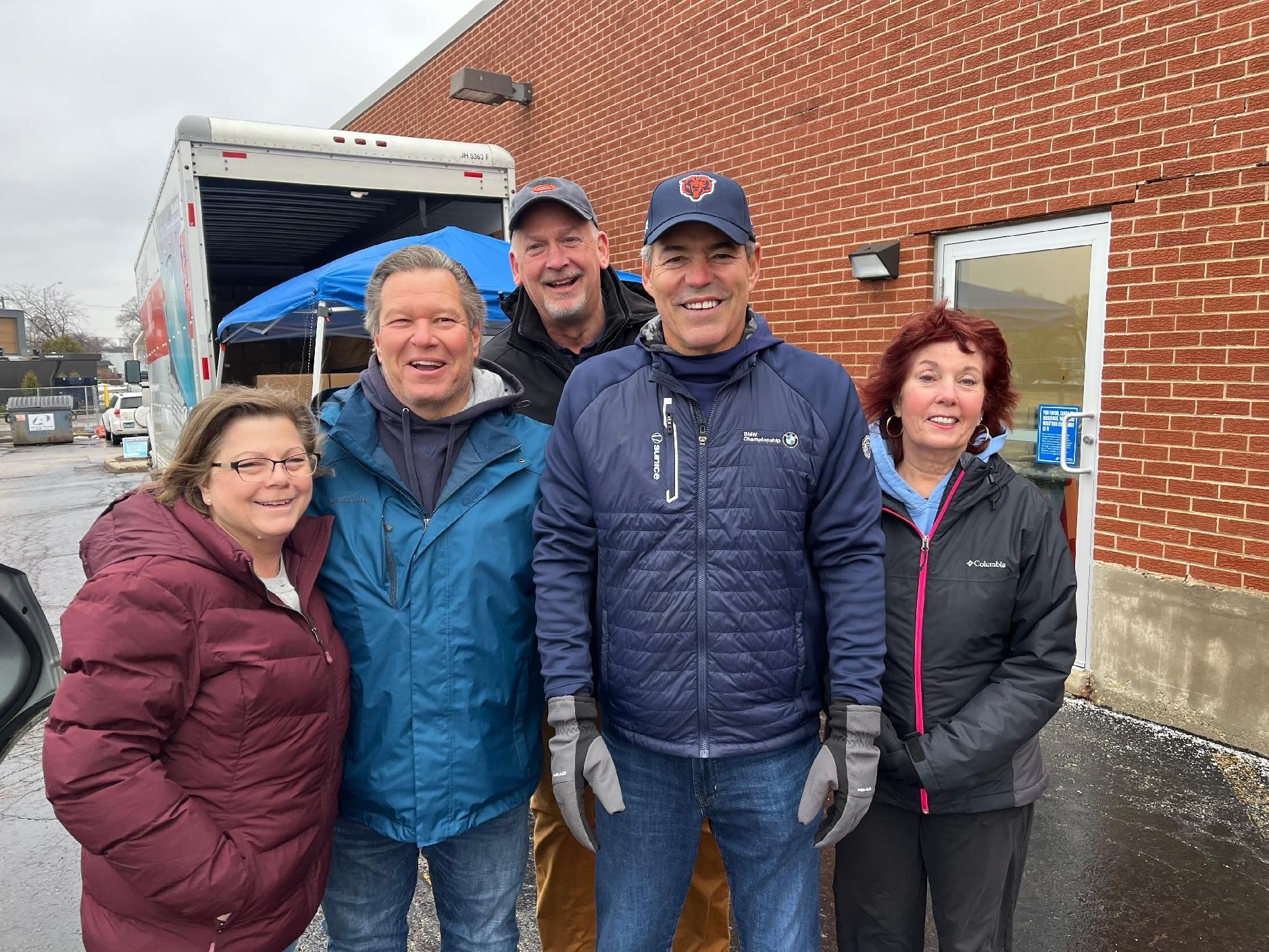 Five people smiling outdoors by a brick building on a wet day, wearing winter jackets and hats.