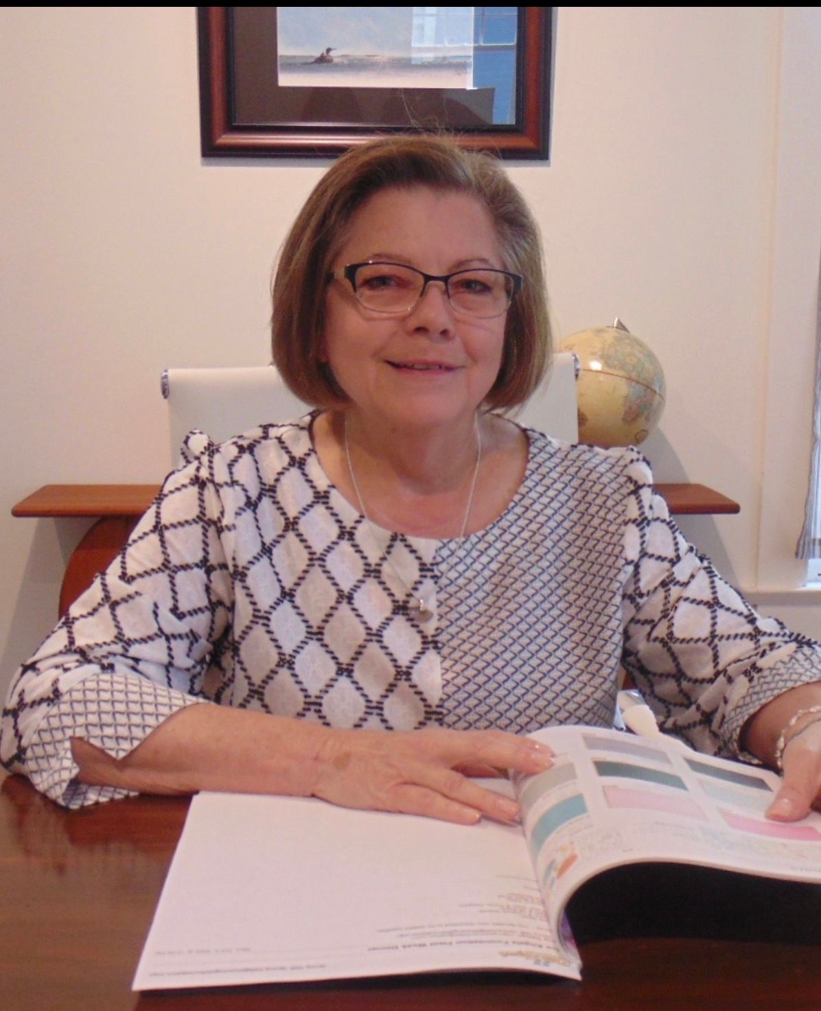 Woman seated at a desk, reading an open book in a bright office setting.