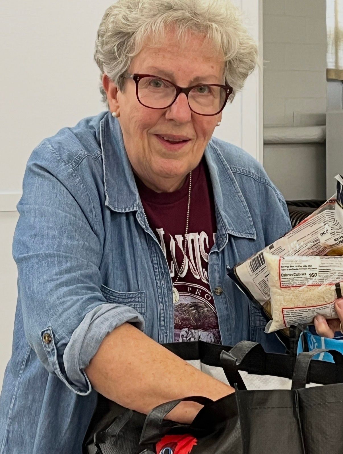 Older woman in denim shirt smiling and holding packaged items at a table in a bright indoor setting