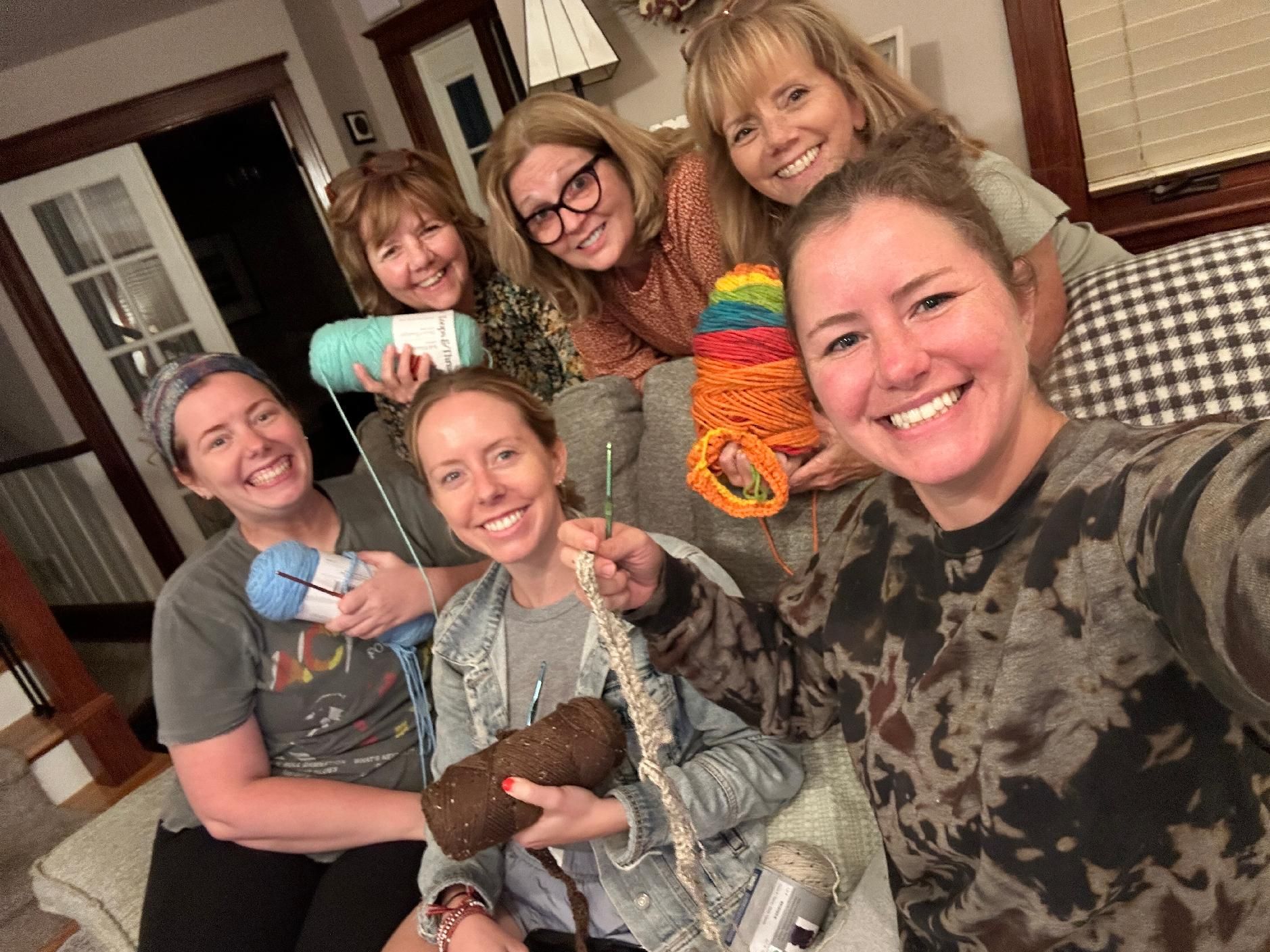 Group selfie of six people indoors, smiling and holding colorful yarn crafts on a couch