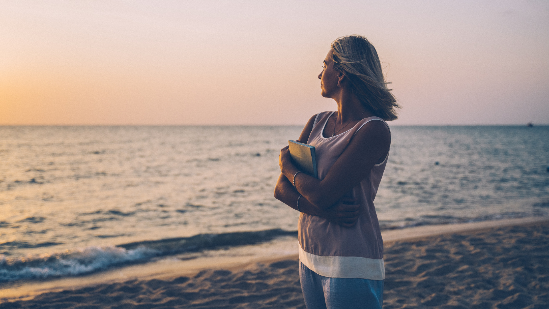 A woman is standing on a beach holding a book and looking at the ocean.