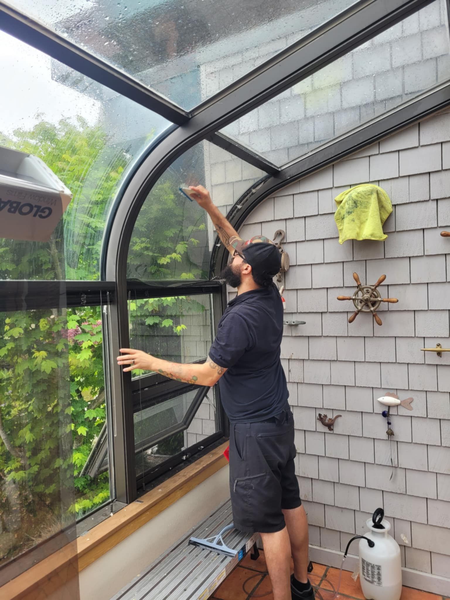 Person cleaning the inside of a glass greenhouse roof with a long-handled tool and cloths nearby