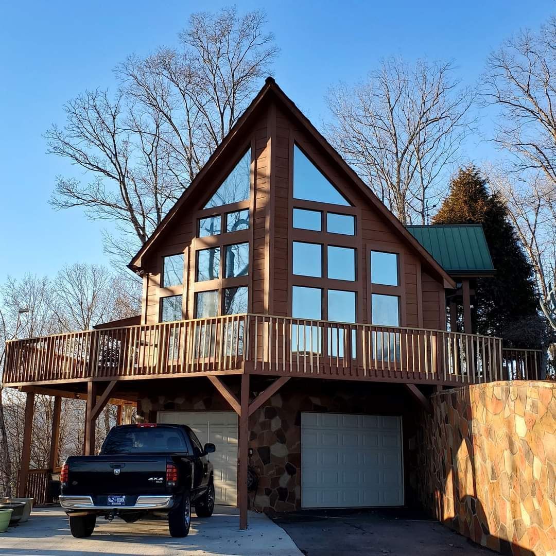 A wooden cabin with large windows and deck, parked black truck below, on a sloped wooded lot.
