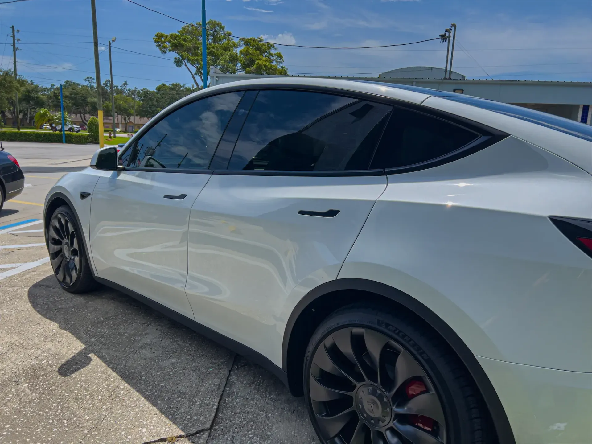 White Tesla Model Y parked on pavement, black rims, tinted windows, bright outdoor setting.