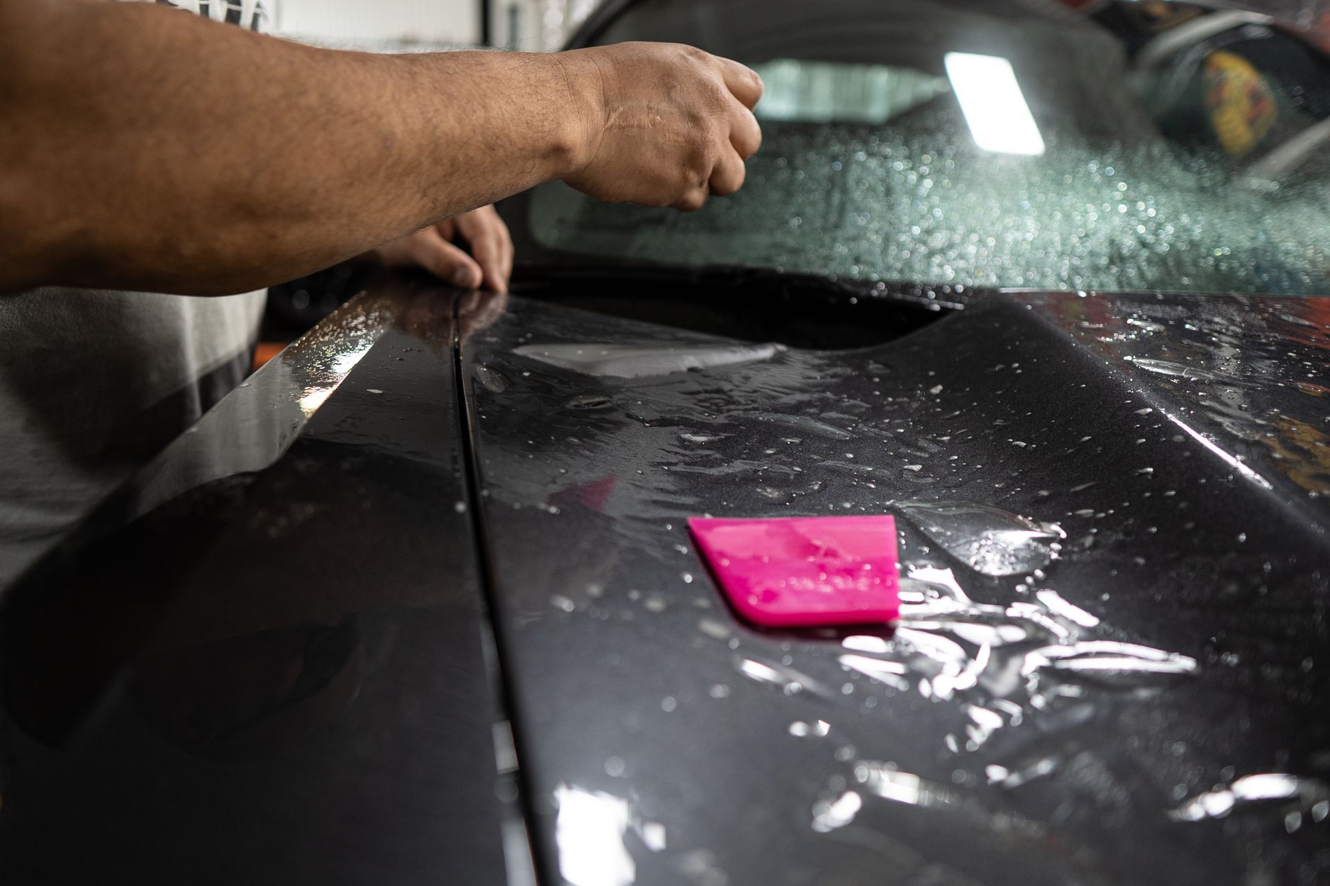 Person installing protective film on a car's hood, using a pink squeegee in a garage.