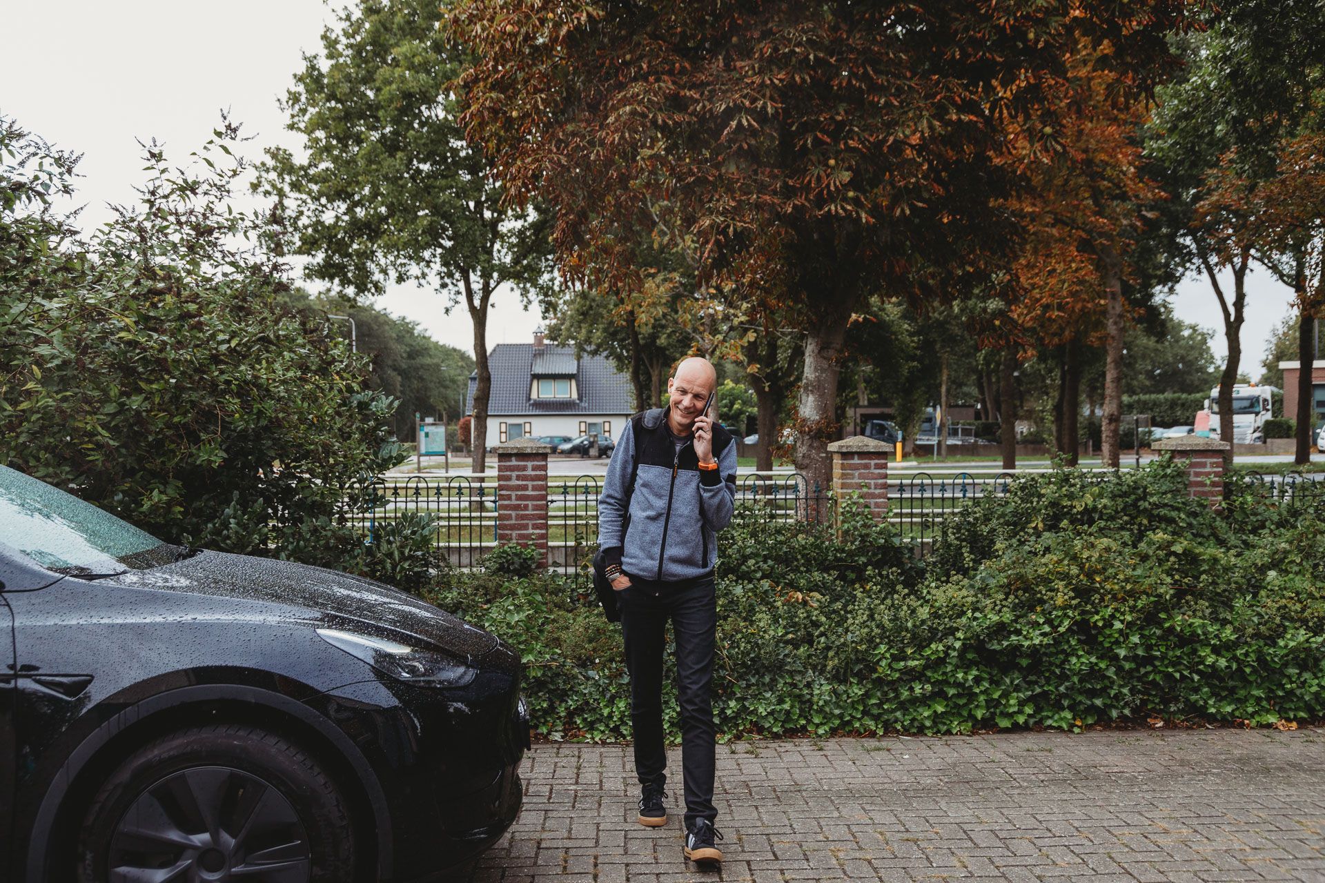 Een persoon staat op een stenen pad naast een zwarte auto en houdt een kind vast in een park met bomen.
