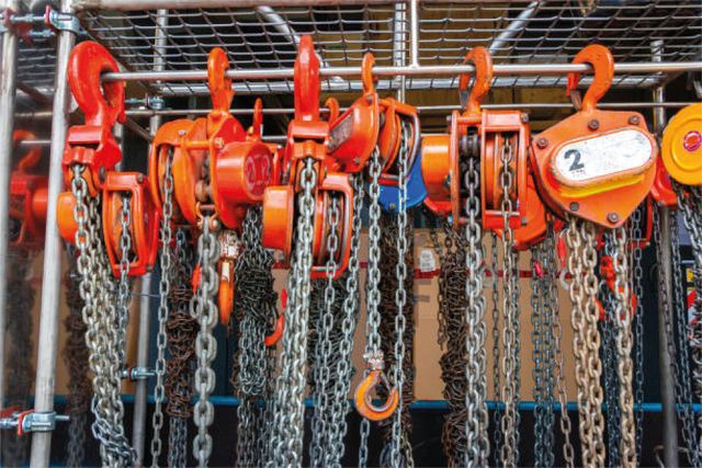Oranje kettingtakels en haken hangen in rijen aan een metalen rek in een industriële omgeving.